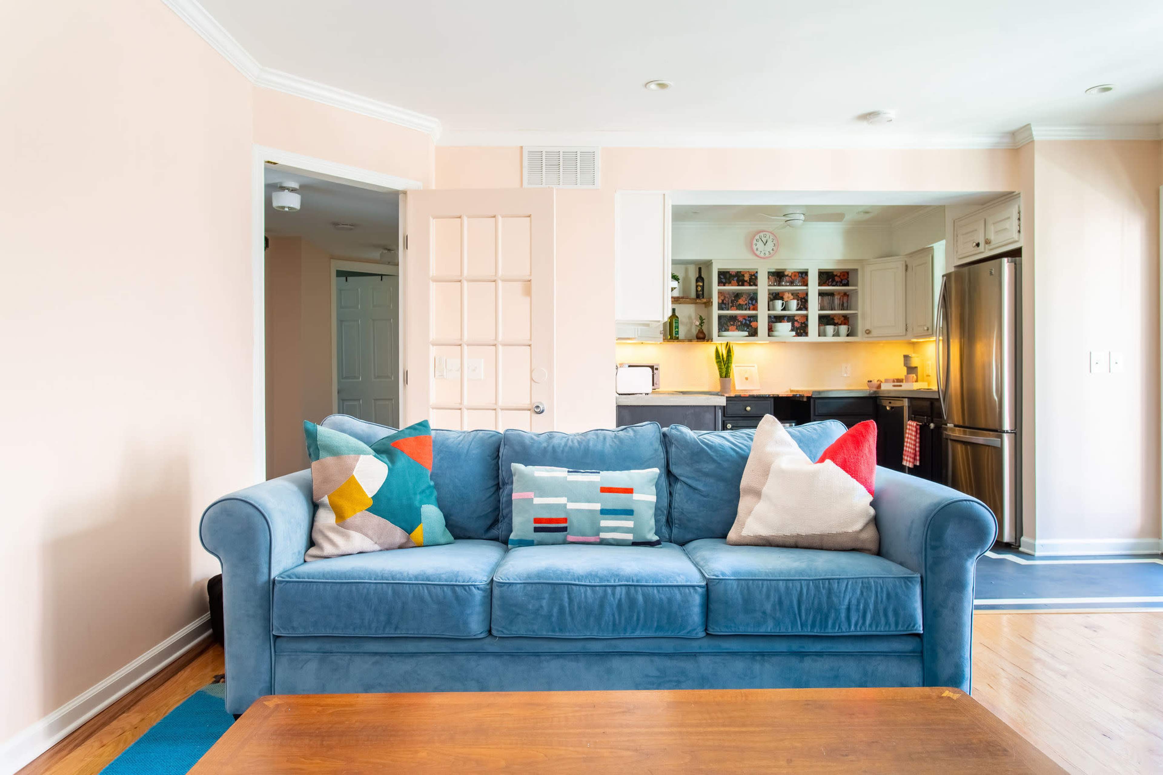 A blue sofa with patterned pillows is positioned in a bright living area that opens into a kitchen with wooden cabinets and appliances.