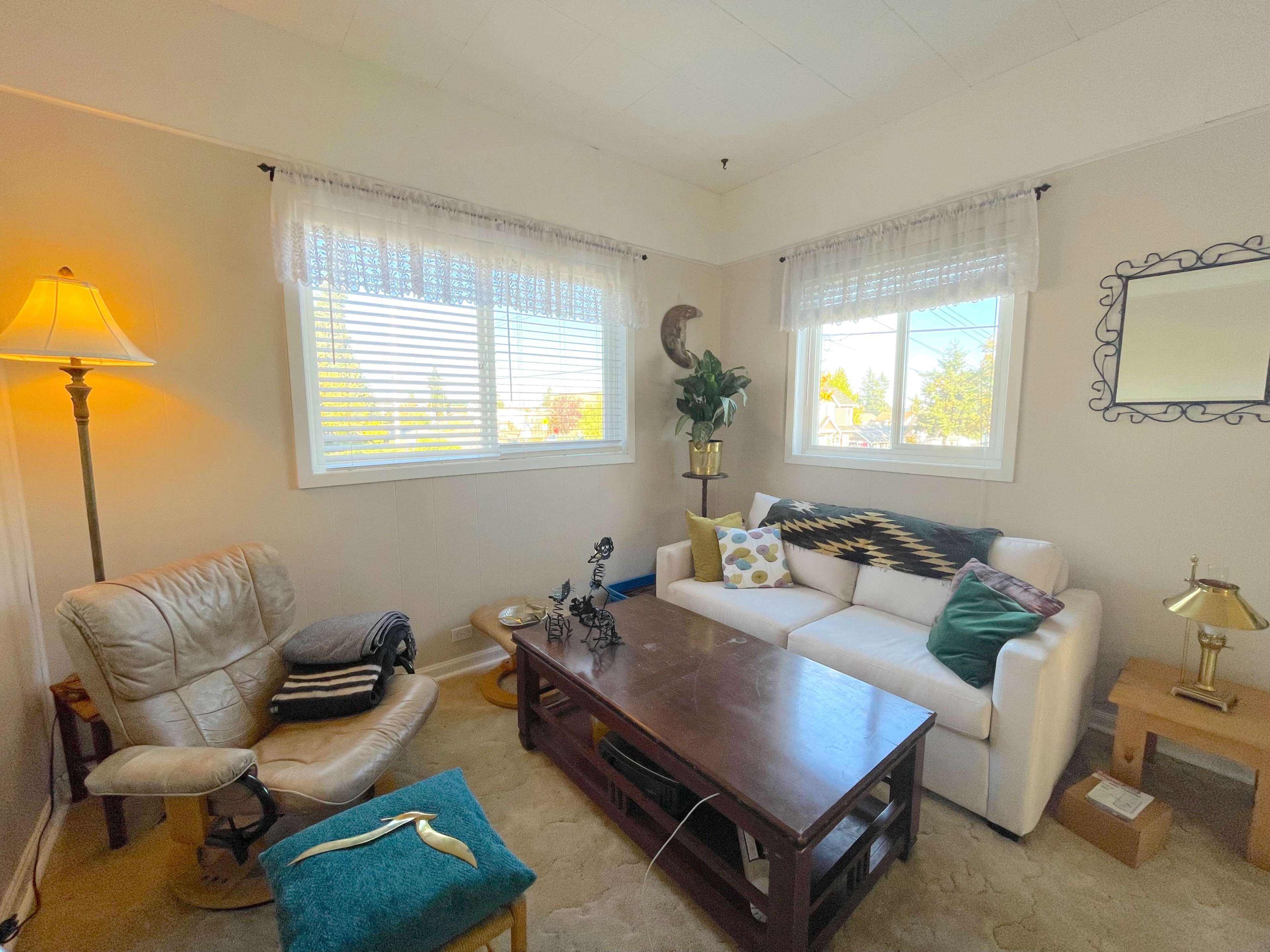 A cozy living room with a white sofa, a wooden coffee table, a comfortable armchair, and two windows allowing natural light to enter.