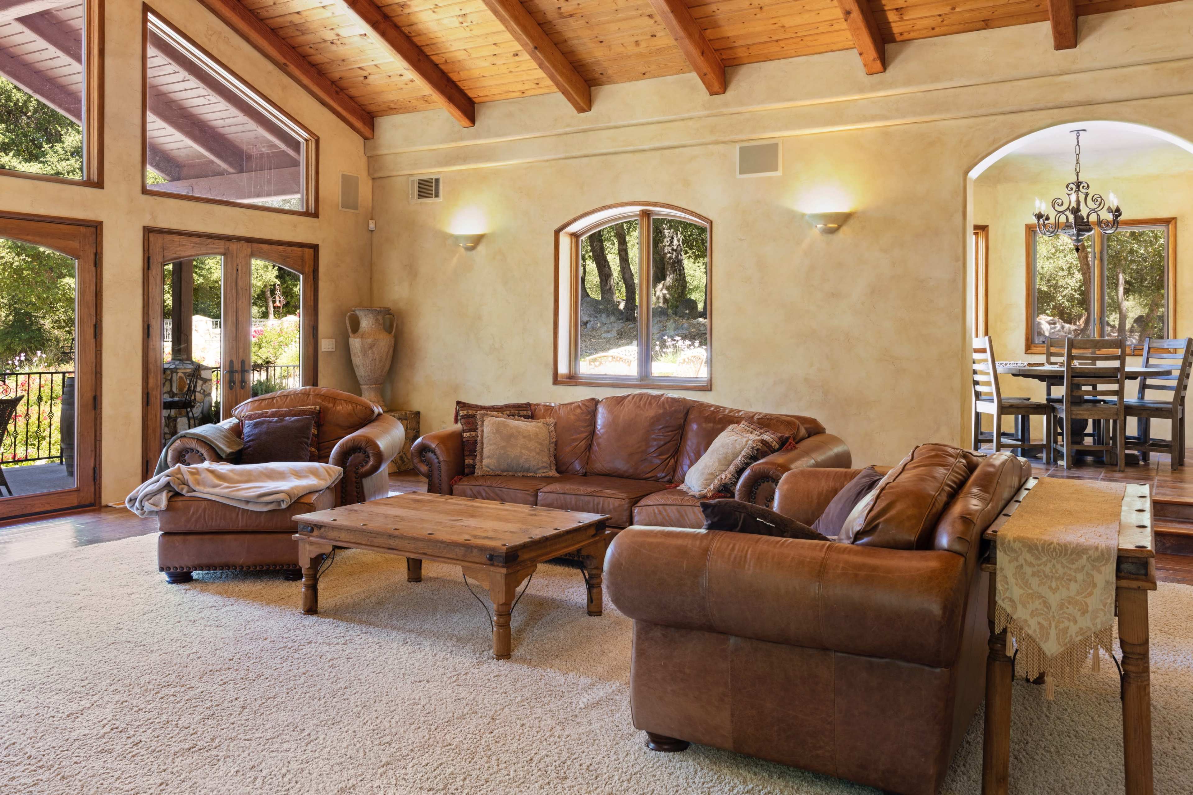 The image shows a cozy living room with leather sofas, a wooden coffee table, and large windows allowing natural light to enter.