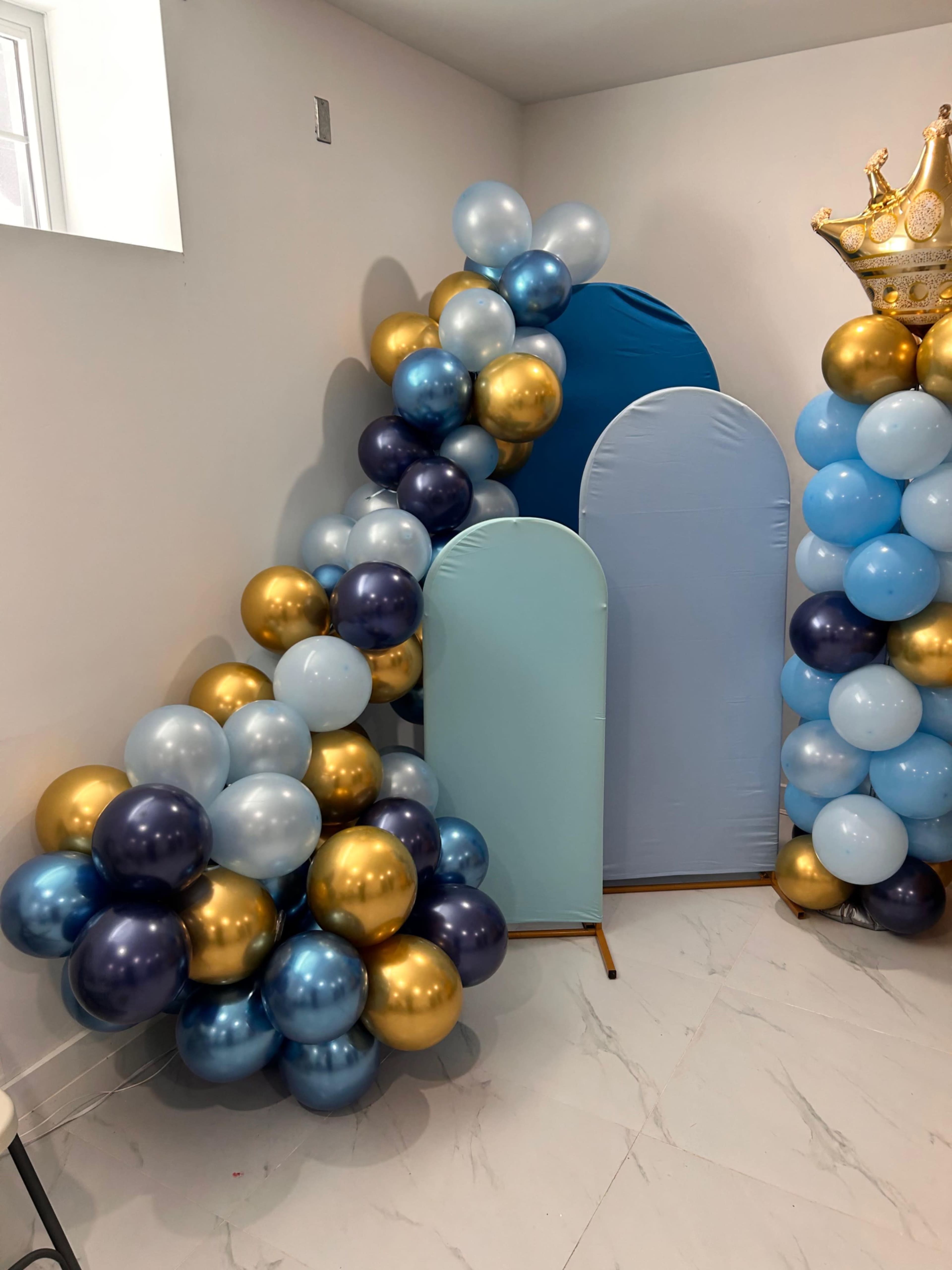 The image shows a decorative corner featuring clusters of balloons in blue, gold, and silver, along with two blue arch-shaped panels and a gold crown atop one side.