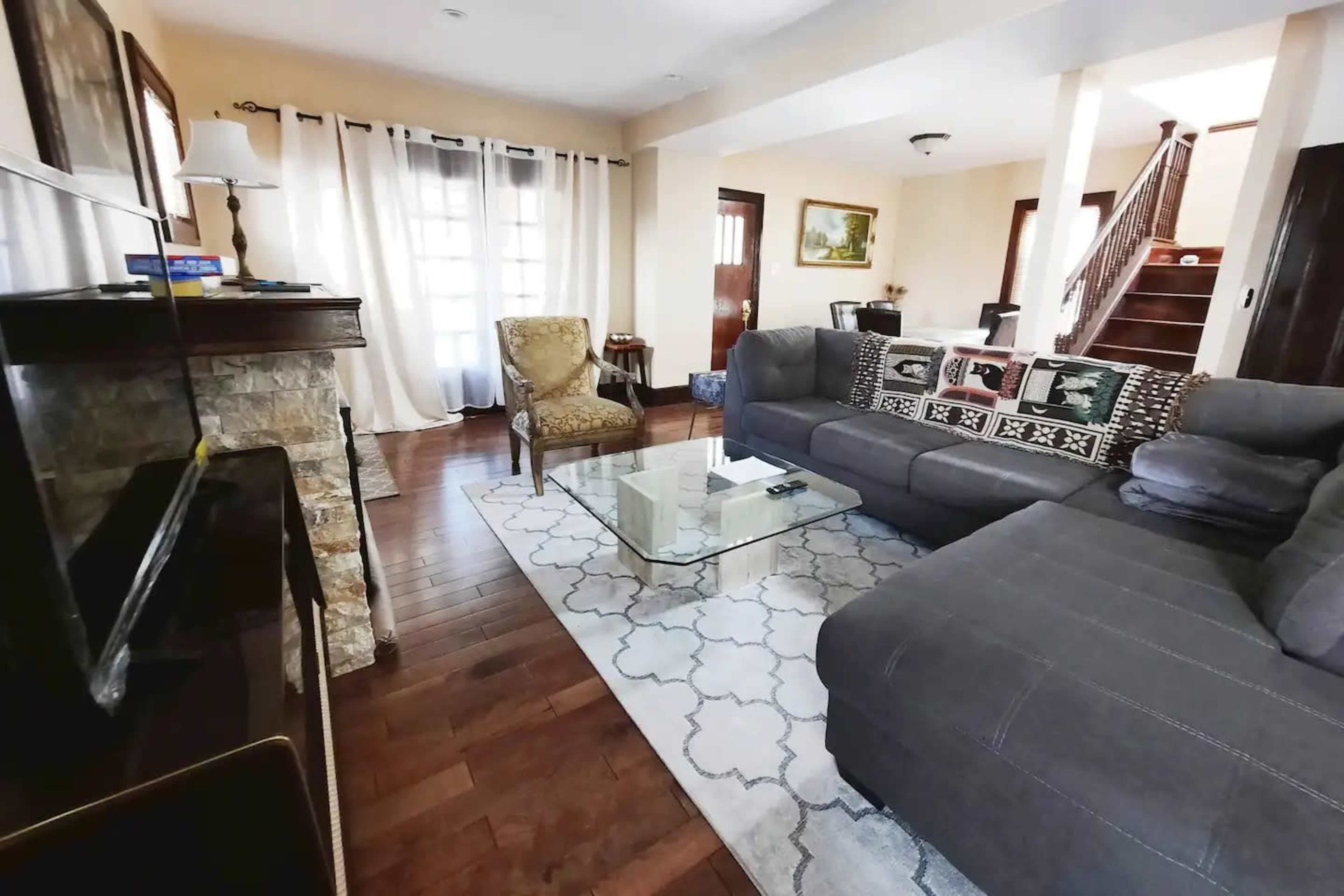 A living room with a gray sectional sofa, a glass coffee table, a patterned rug, and a chair, featuring warm lighting and wooden floors.