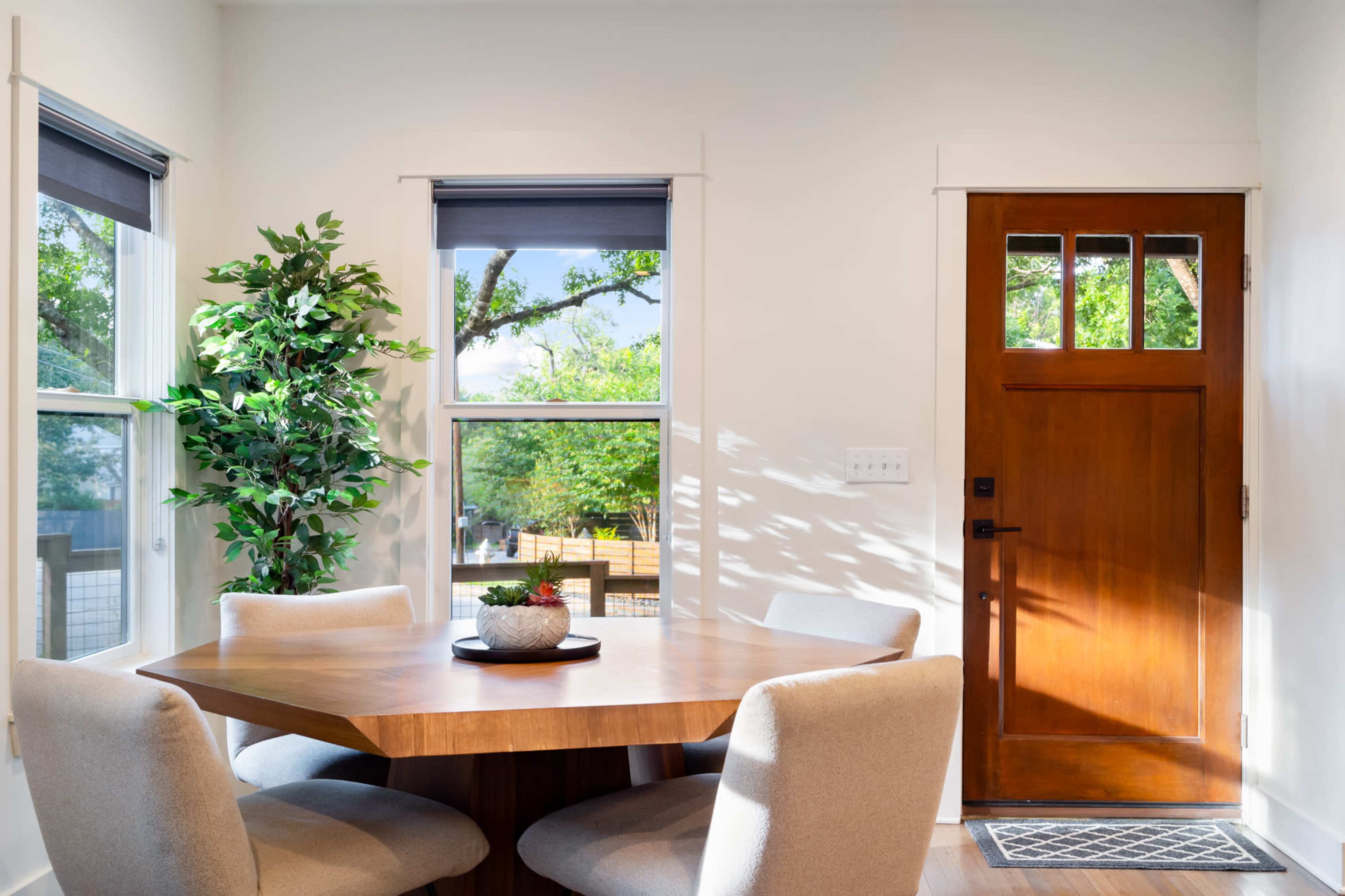 A wooden dining table surrounded by four light-colored chairs is positioned near a door and windows that provide natural light and a view of outdoor greenery.