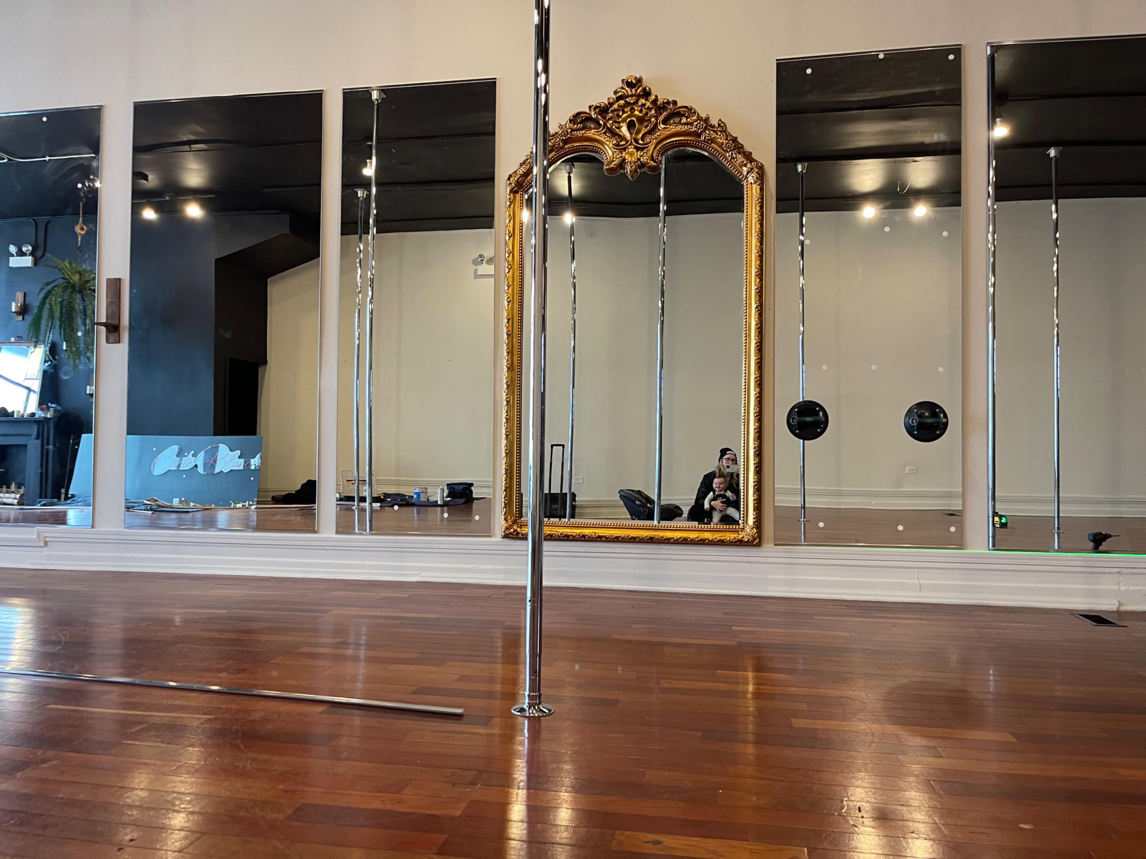 A spacious room with polished wooden floors, featuring large mirrors, a decorative golden mirror, and a pole in the center.