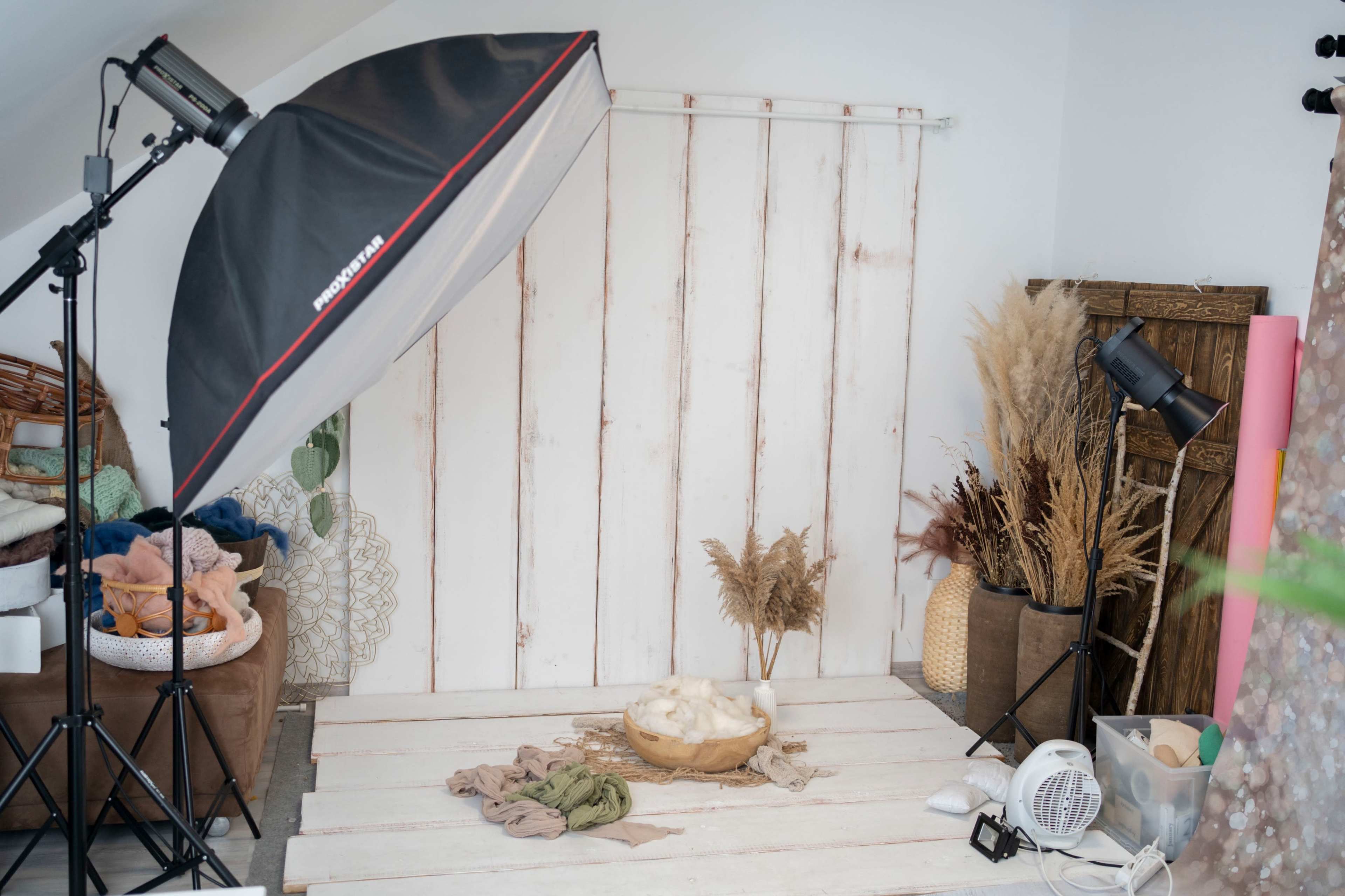 The image shows a photography setup featuring a large softbox light and various props including dried plants, fabrics, and a wooden bowl on a whitewashed wooden surface.