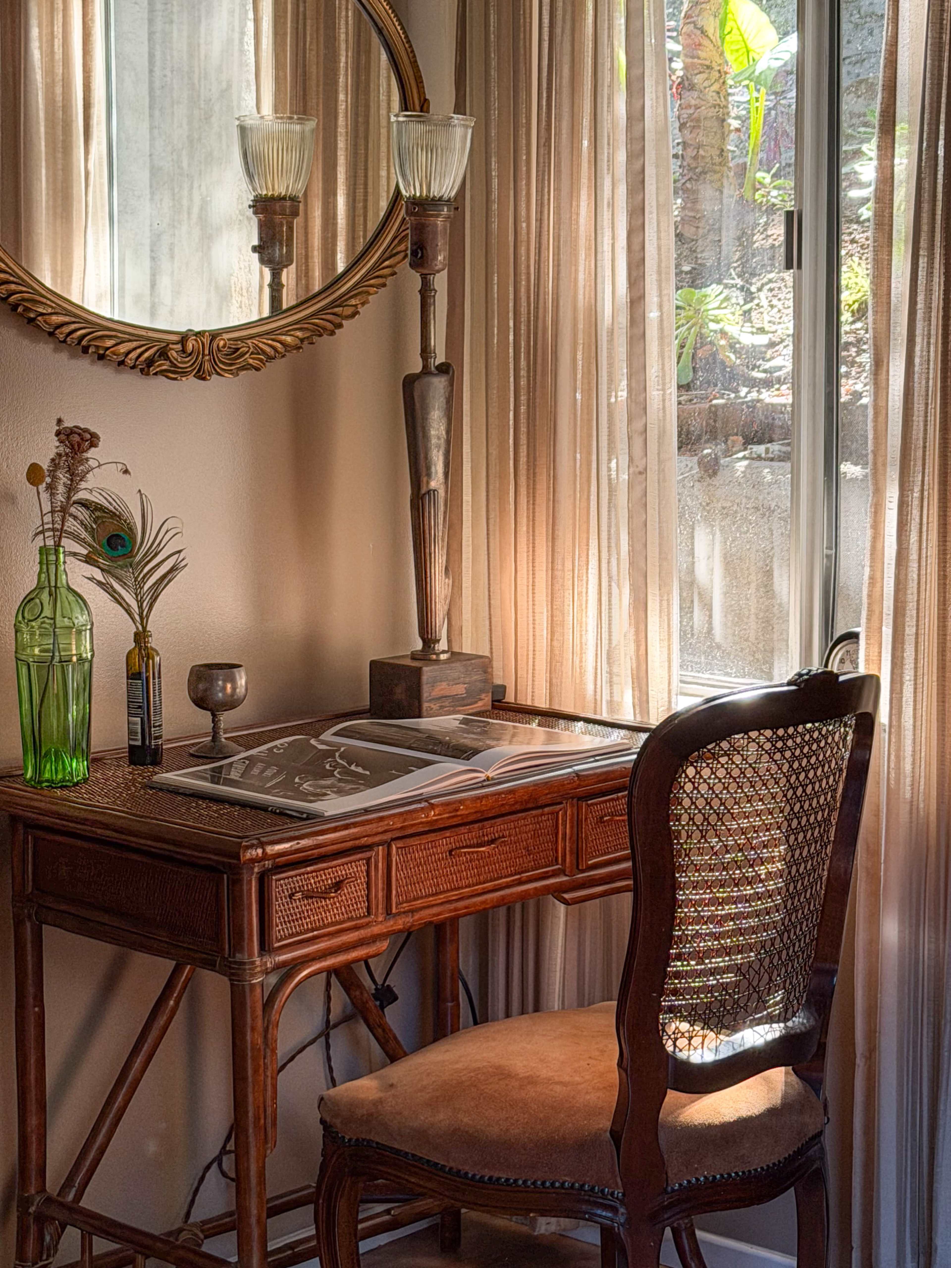A vintage wooden desk with a chair, a round mirror above it, and a soft light filtering through sheer curtains beside a window.