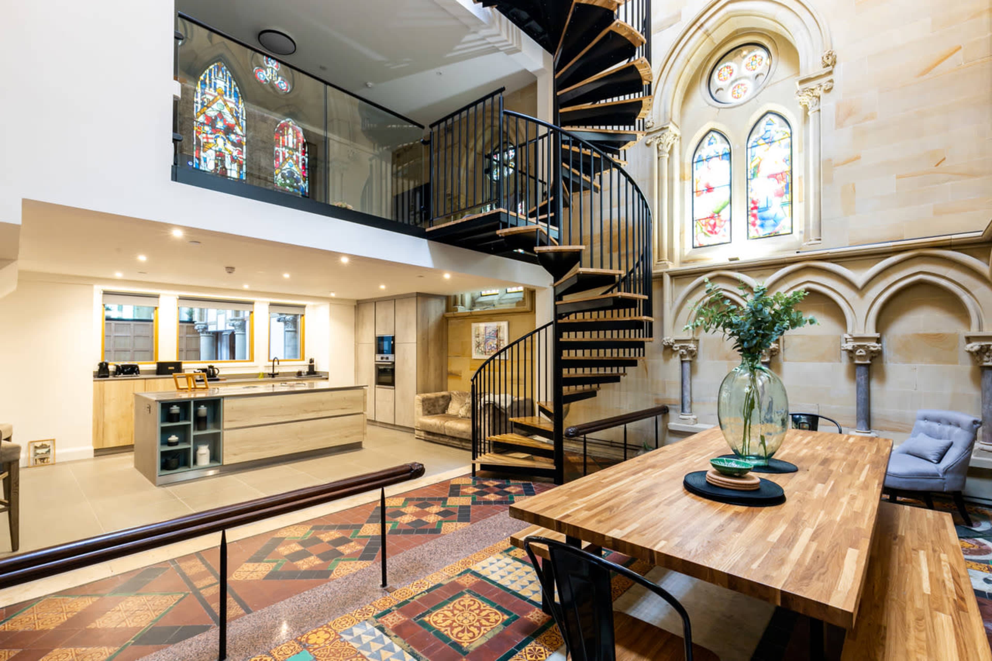 A modern interior space featuring a spiral staircase, a kitchen area, and a dining table surrounded by colorful tiled flooring and large stained glass windows.