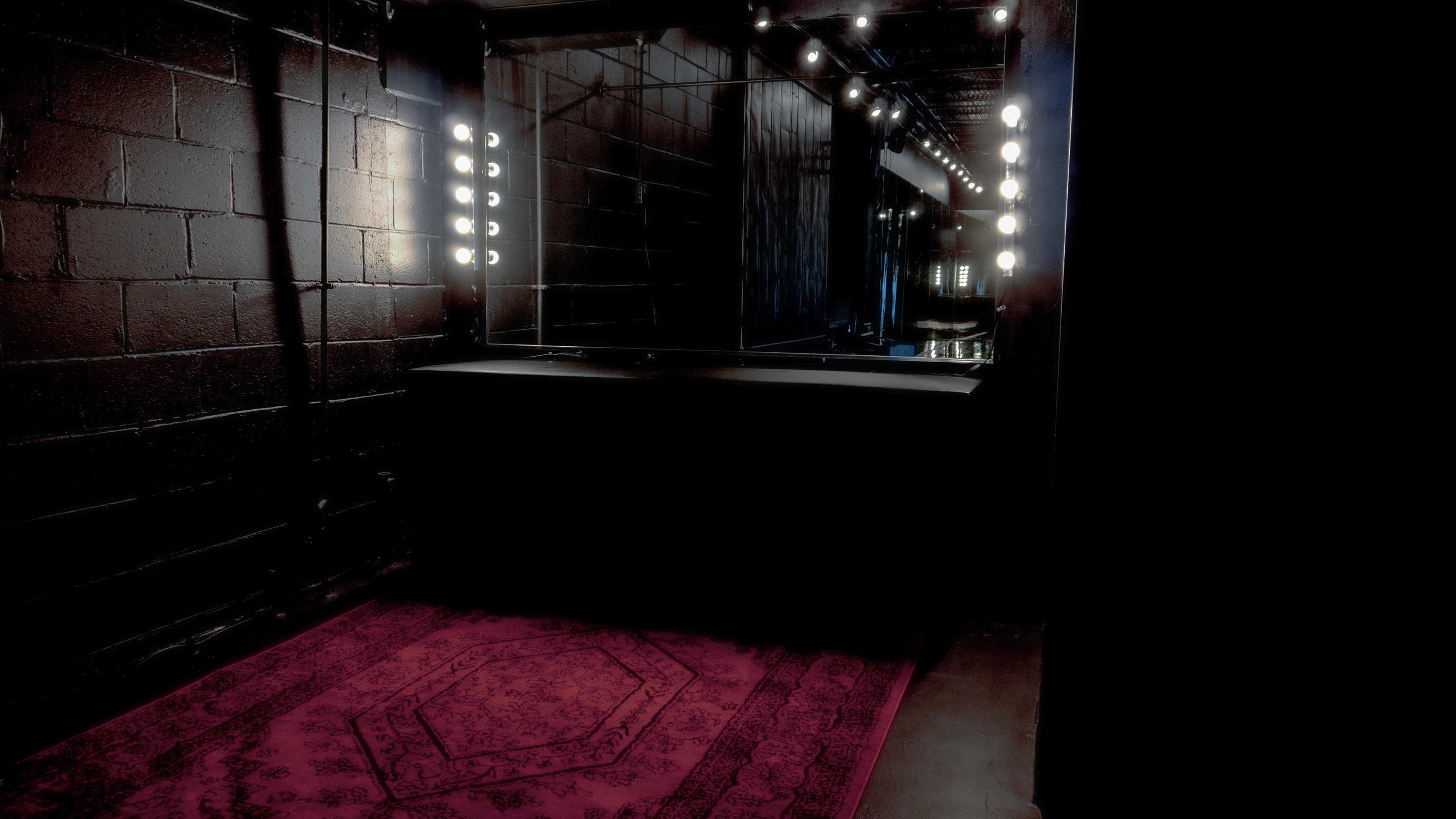 The image shows a dimly lit room featuring a large mirror illuminated by lights, with a dark wall and a red carpet on the floor.