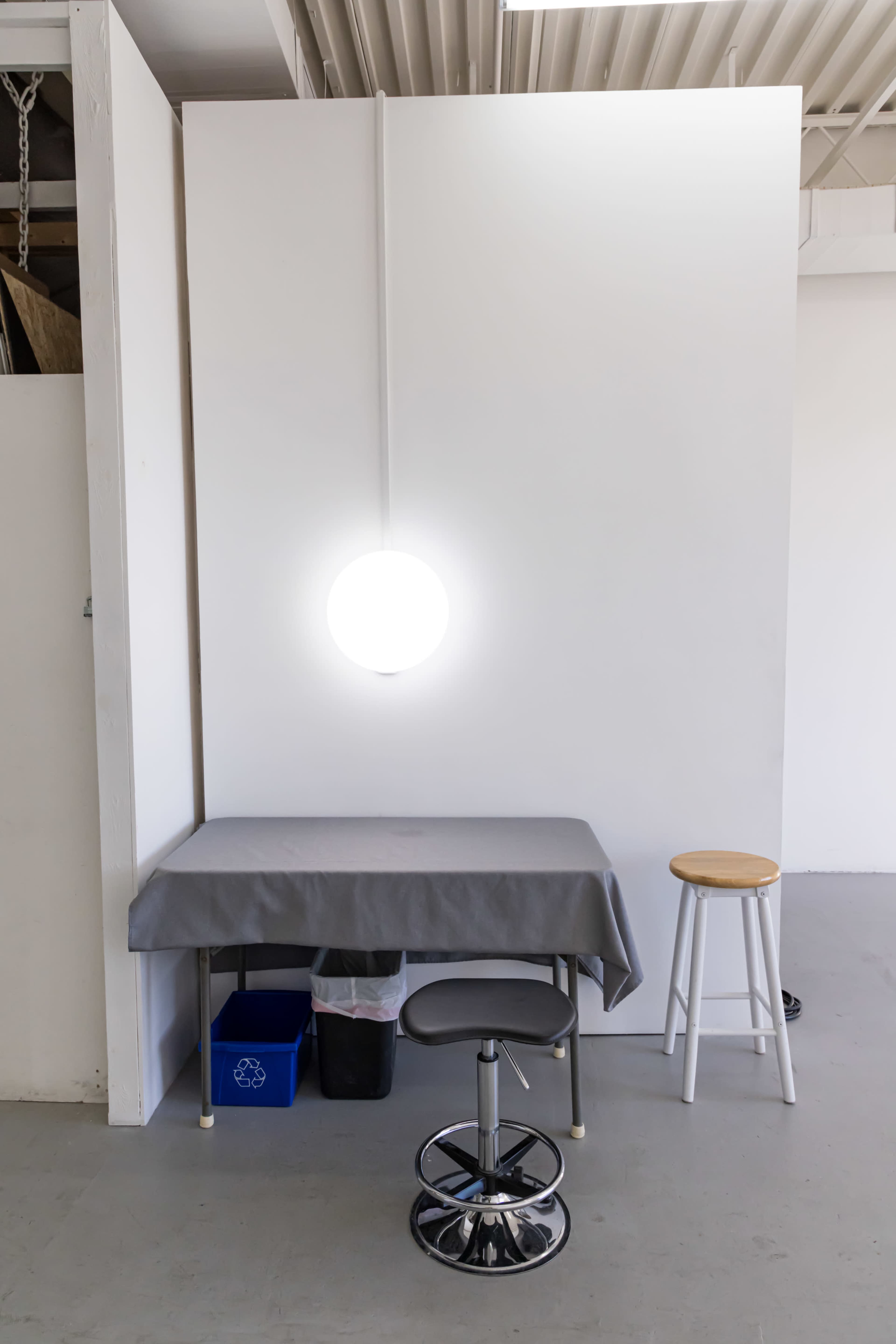 A simple workspace features a table covered with a gray cloth, a stool, a recycling bin, and a round hanging light above.