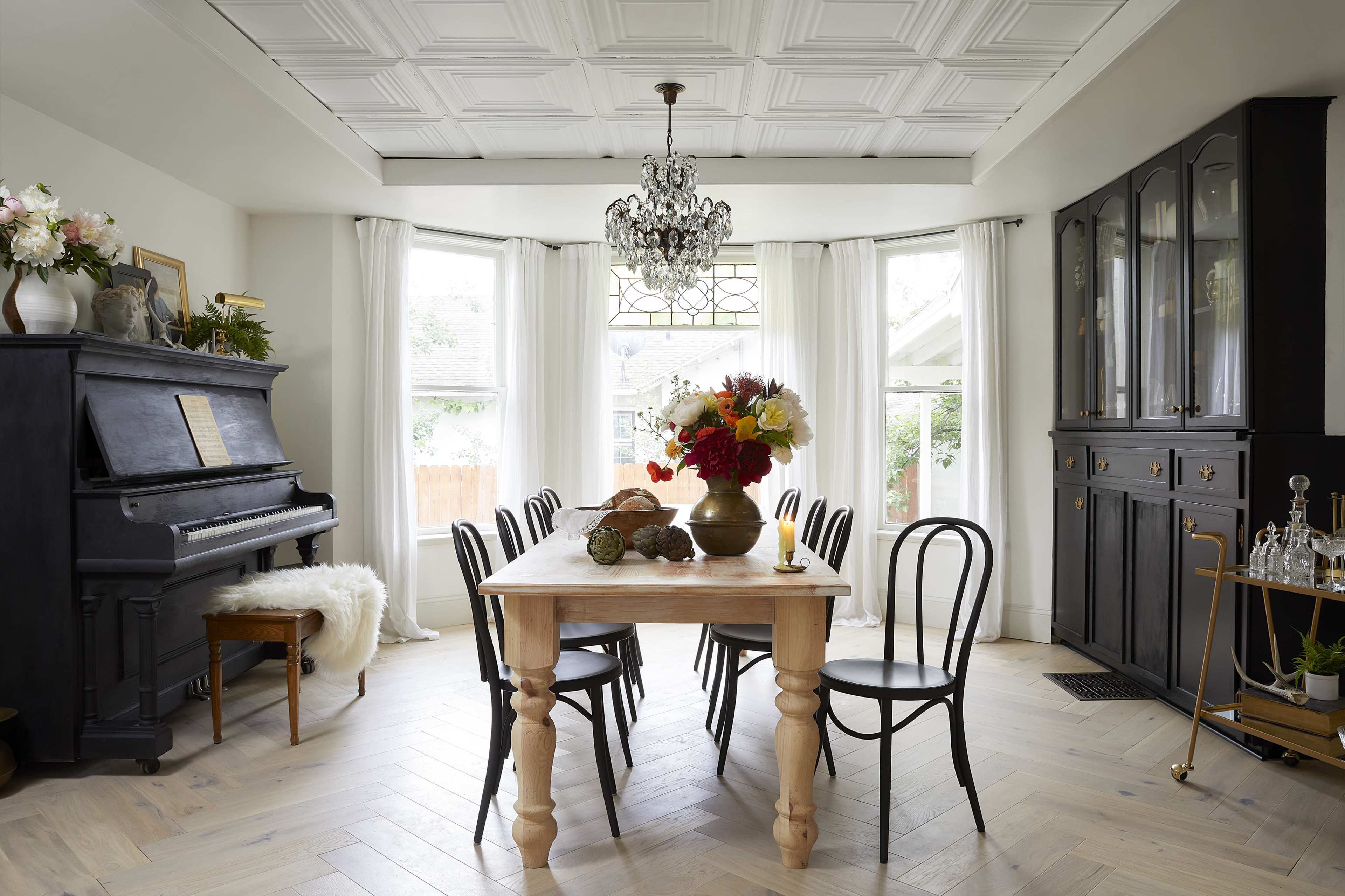 A dining room features a wooden table surrounded by black chairs, with a piano in the corner and a large window that provides natural light.