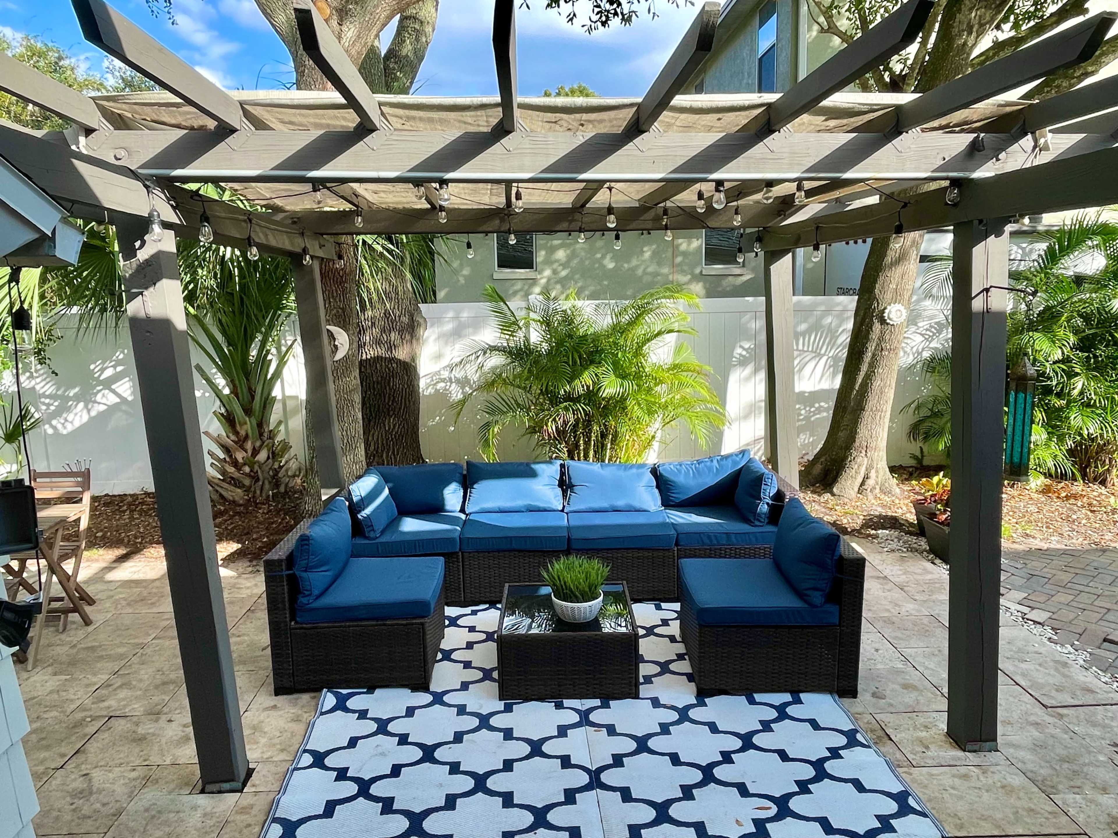 A shaded outdoor seating area features a sectional sofa with blue cushions, a coffee table, and a patterned rug surrounded by greenery and a trellis.
