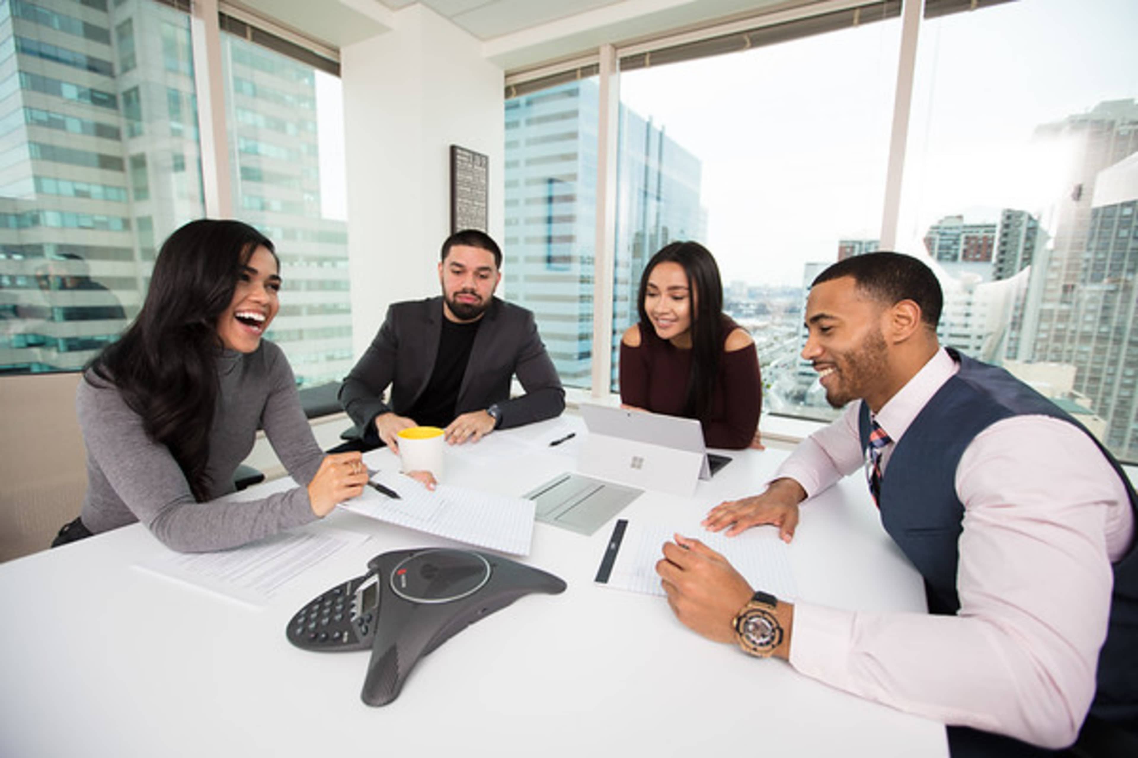 Four professionals are engaged in a meeting around a conference table, with two using laptops and one holding a cup.