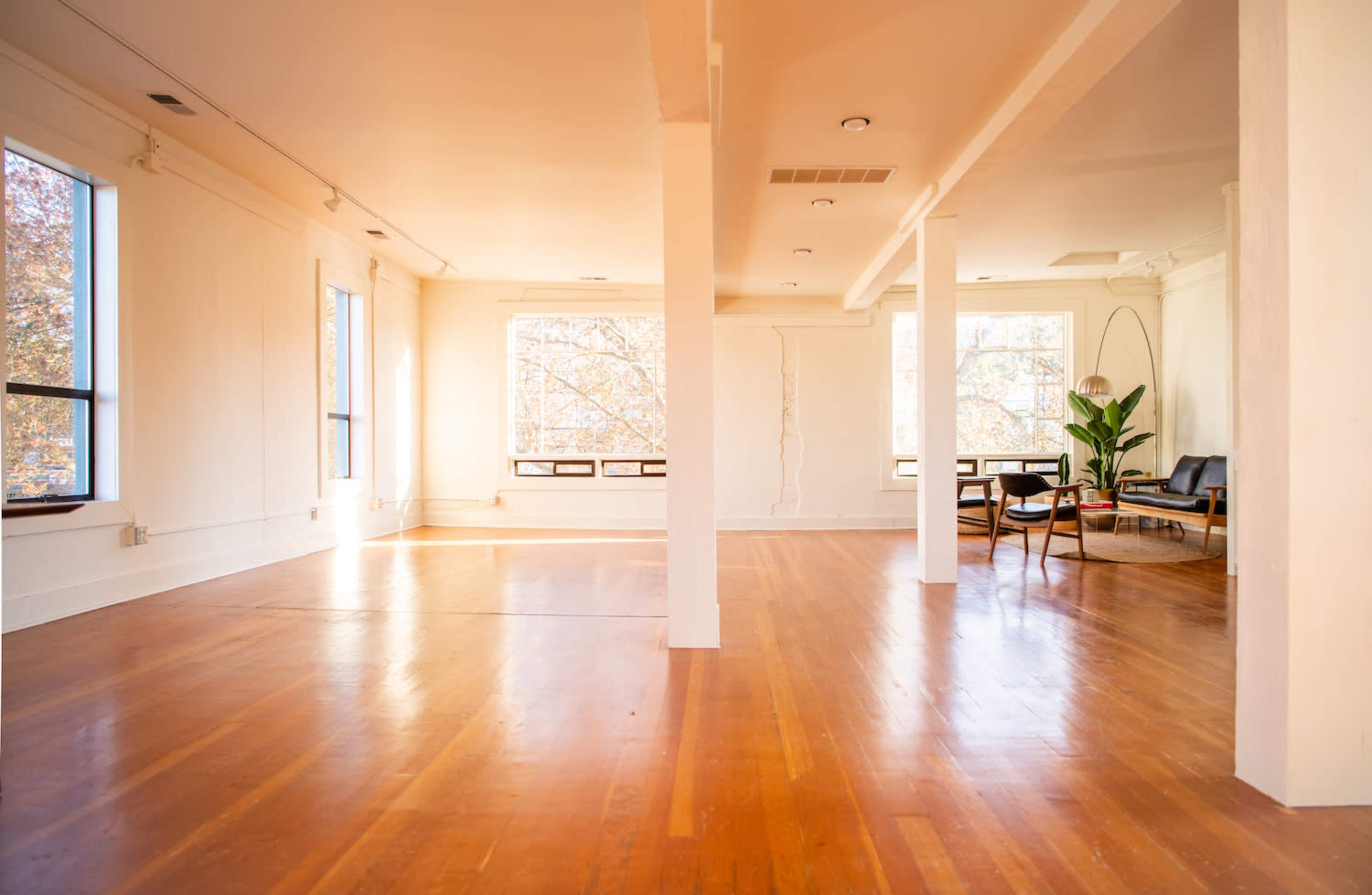 A spacious, empty room with wooden flooring, large windows, and a few pieces of minimal furniture.
