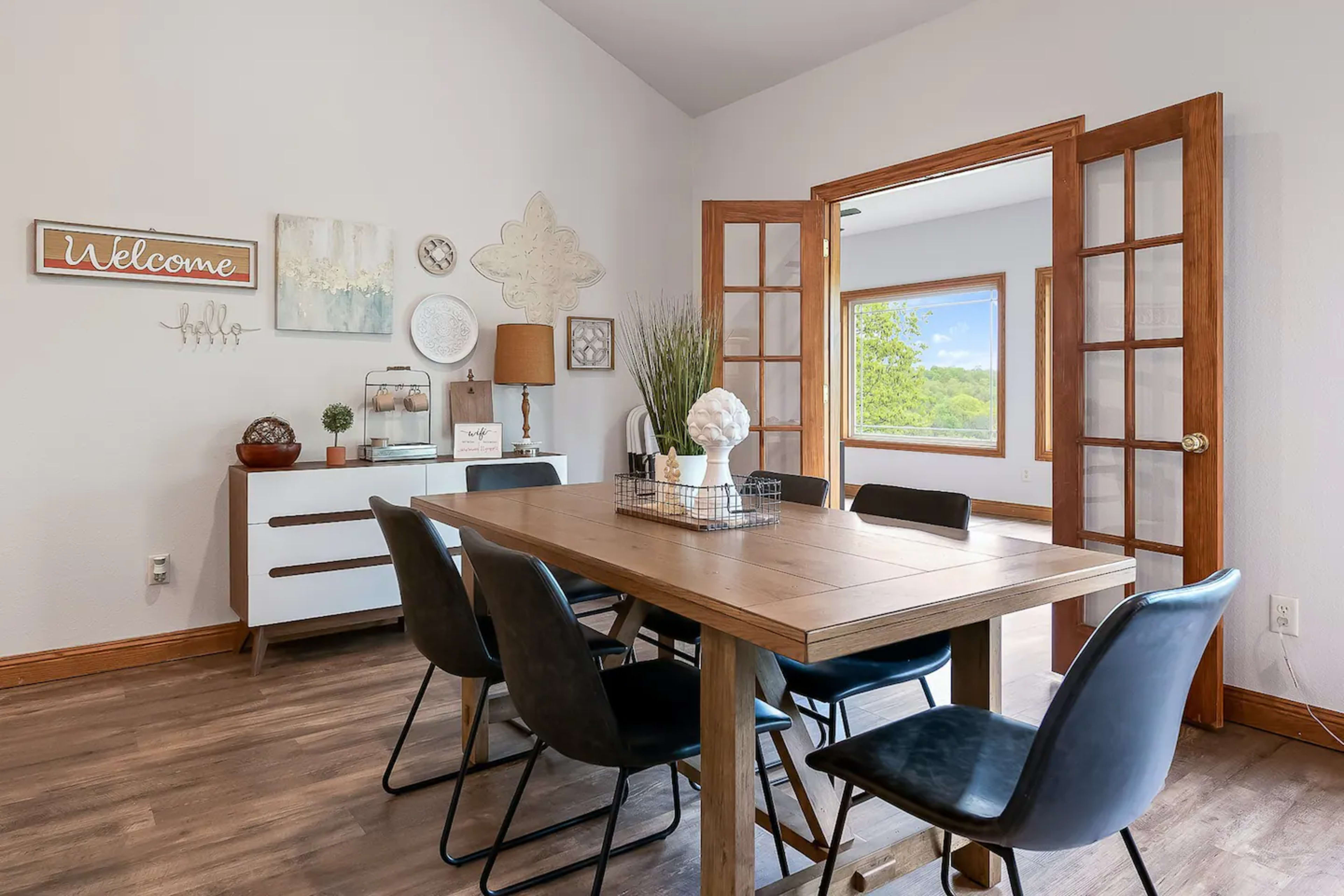 A modern dining area features a wooden table surrounded by black chairs, with a sideboard and decorative wall art in a well-lit room.