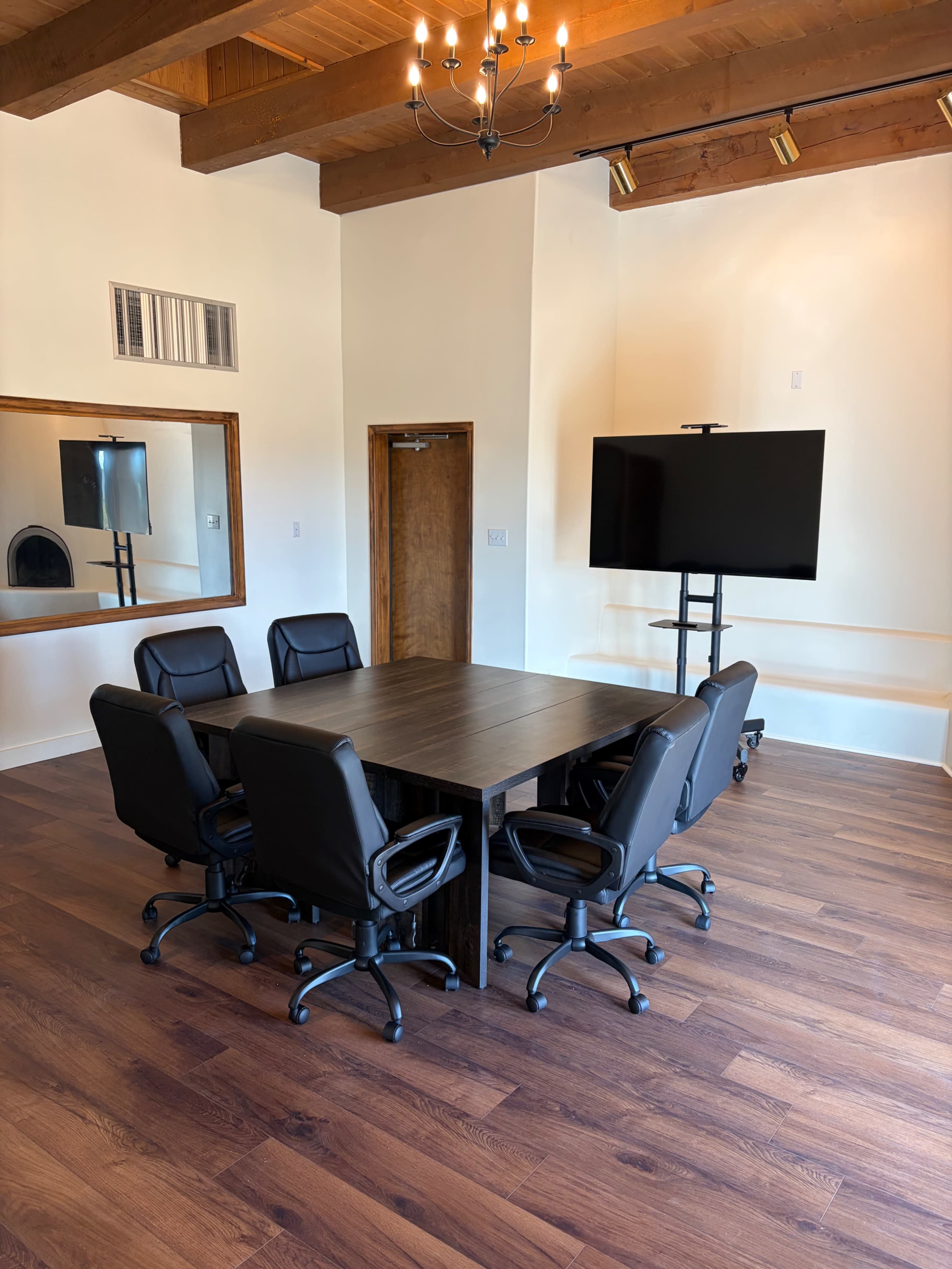 A conference room features a rectangular table surrounded by six black office chairs, with a large flat-screen TV mounted on a stand and exposed wooden beams overhead.