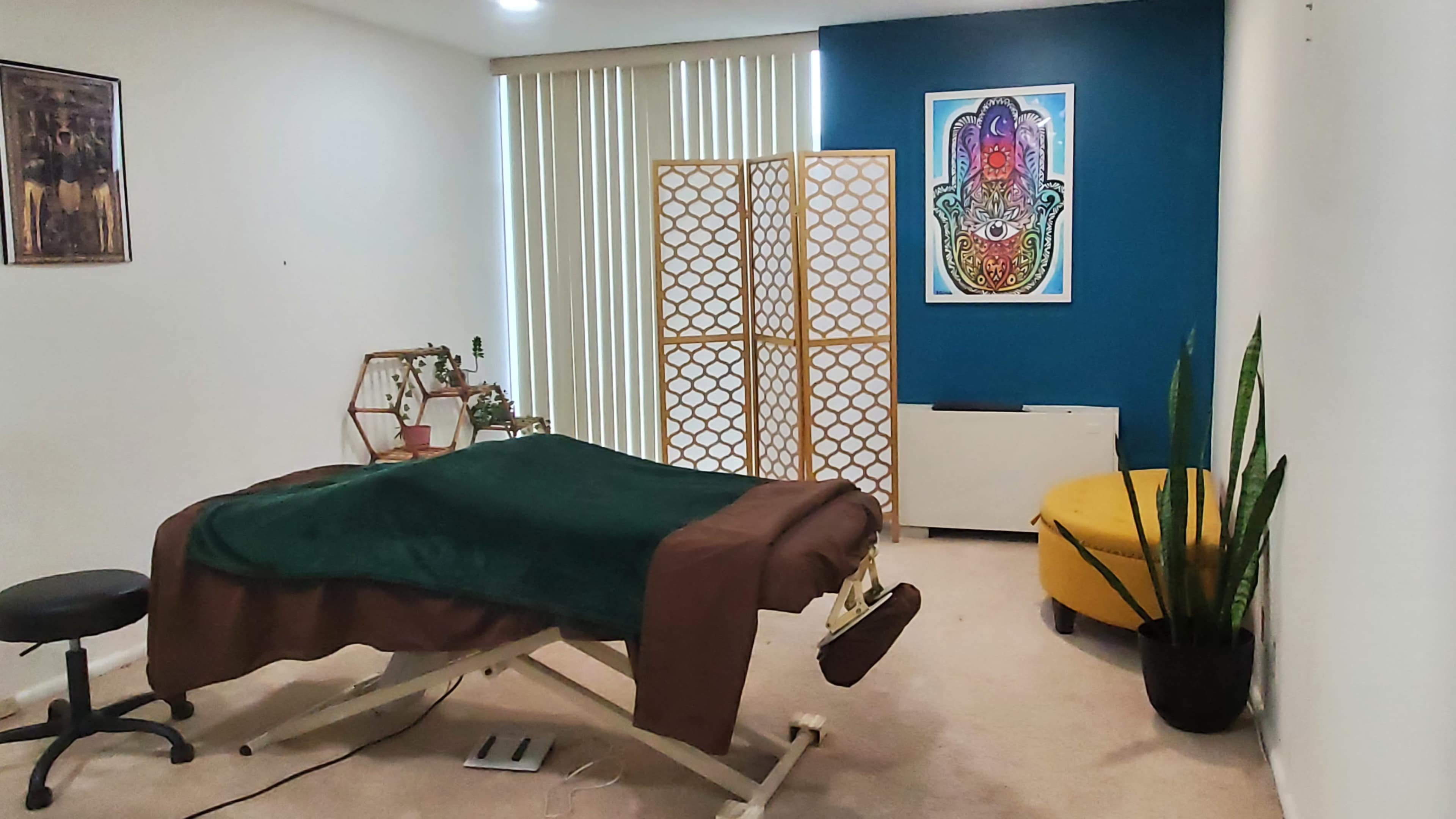 The image depicts a therapy room featuring a treatment table covered with a green blanket, a folding screen, a yellow chair, and wall art.