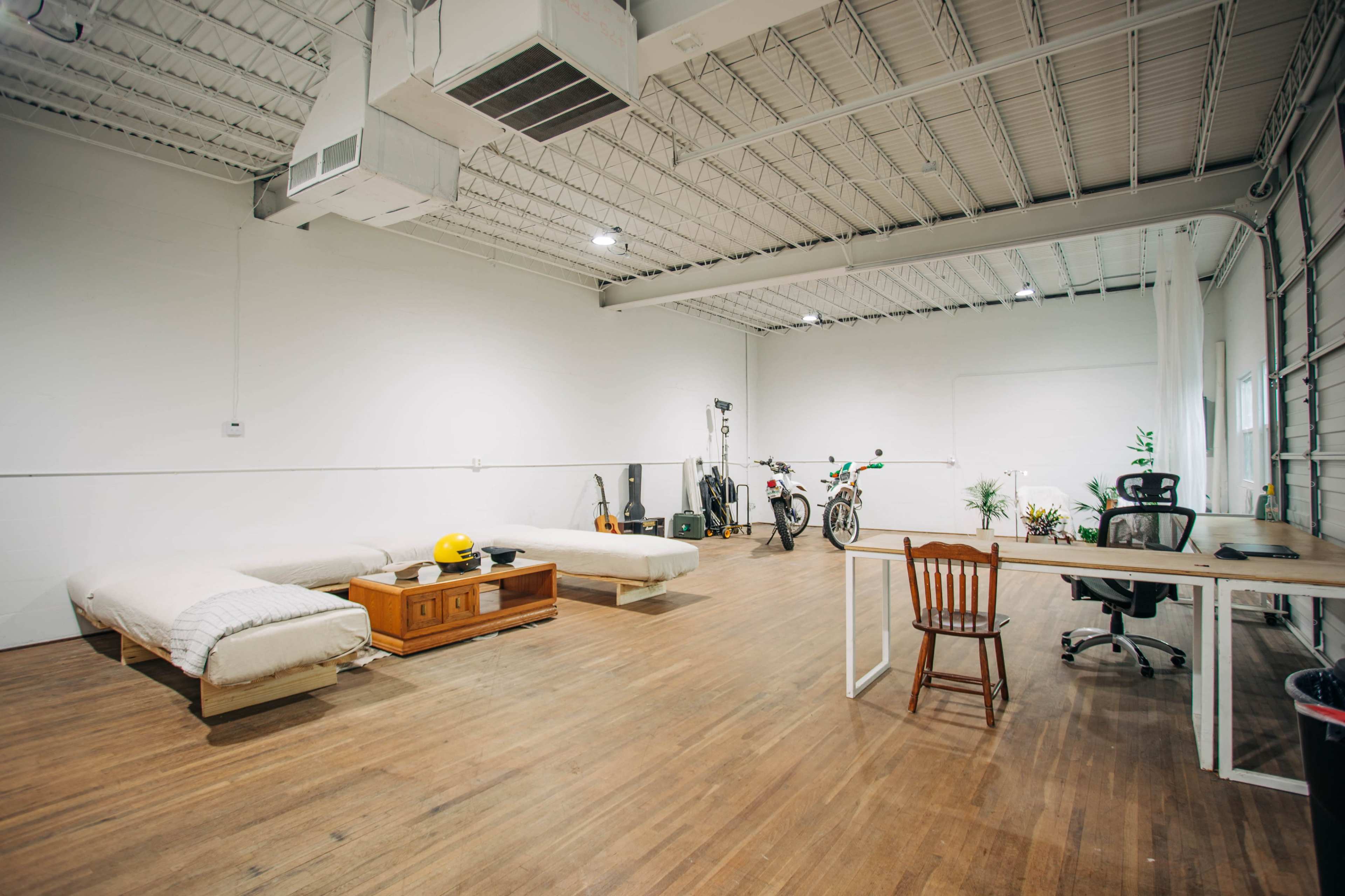 A spacious, minimalistic room with wooden flooring, featuring two beds, a wooden table, a chair, and various items like bicycles and a guitar.