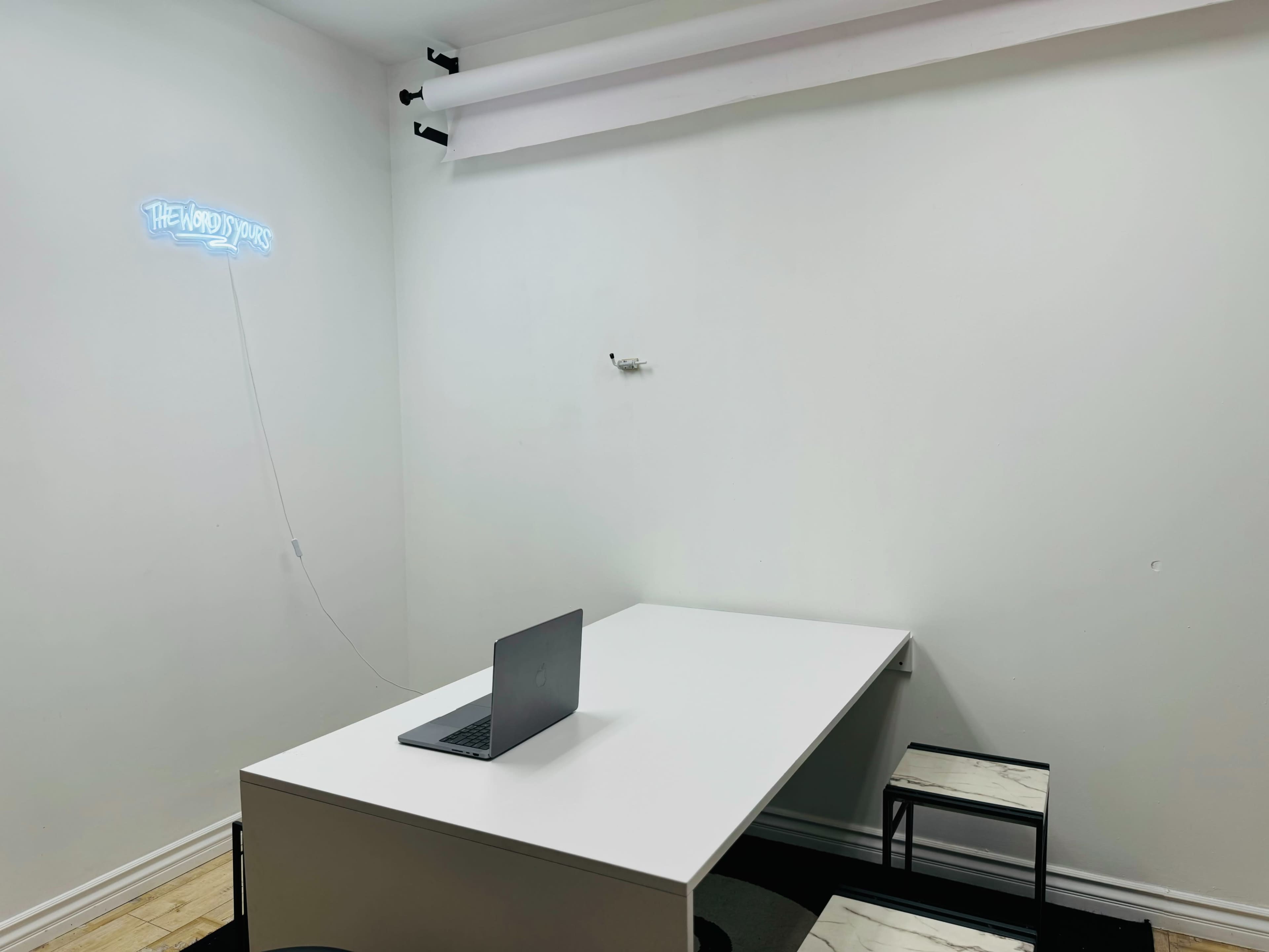 A minimalistic white room features a table with a laptop and two chairs, along with a neon sign on the wall.