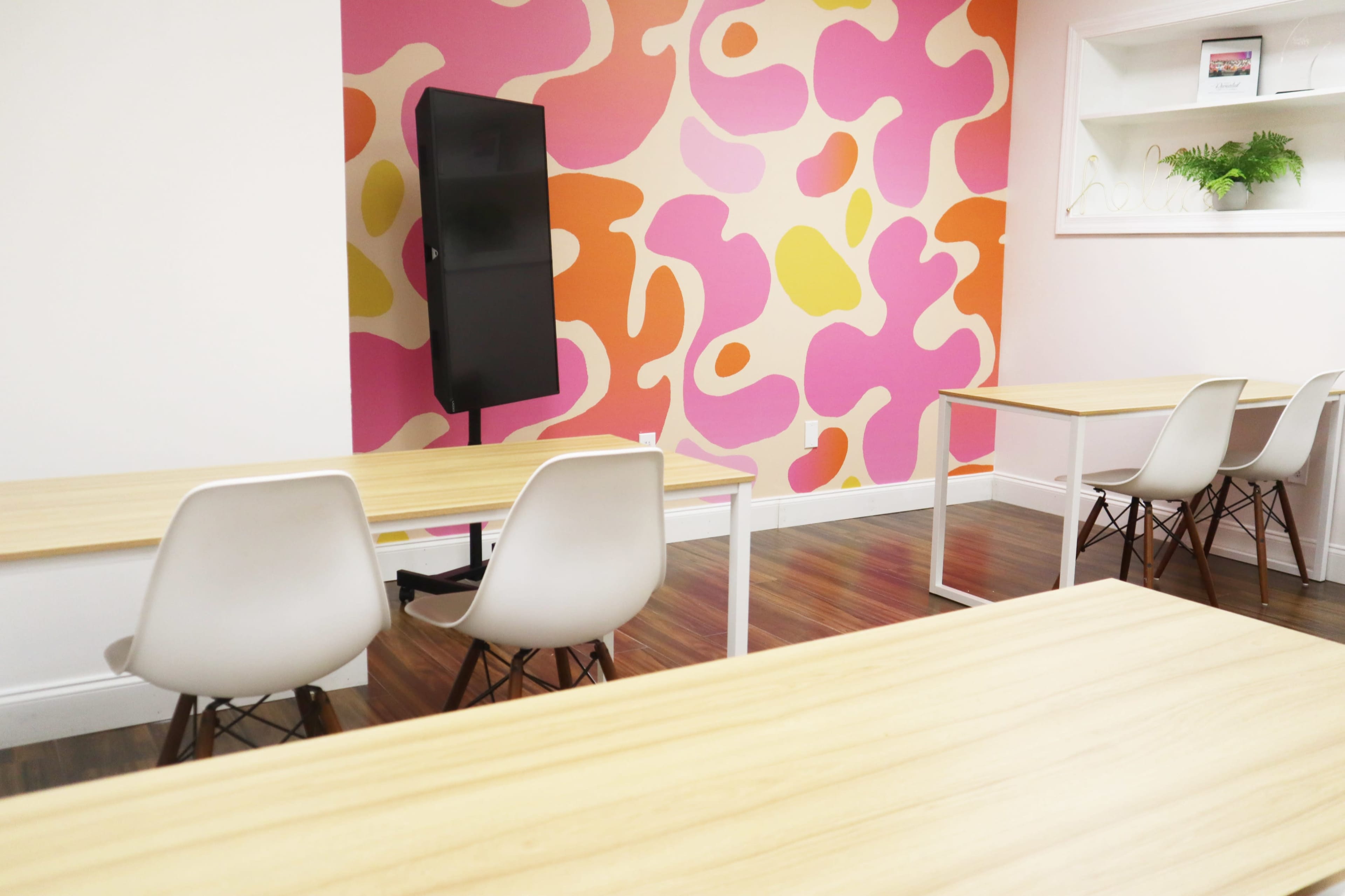 A brightly colored classroom features multiple wooden tables and white chairs facing a black display screen against a vibrant pink and orange patterned wall.
