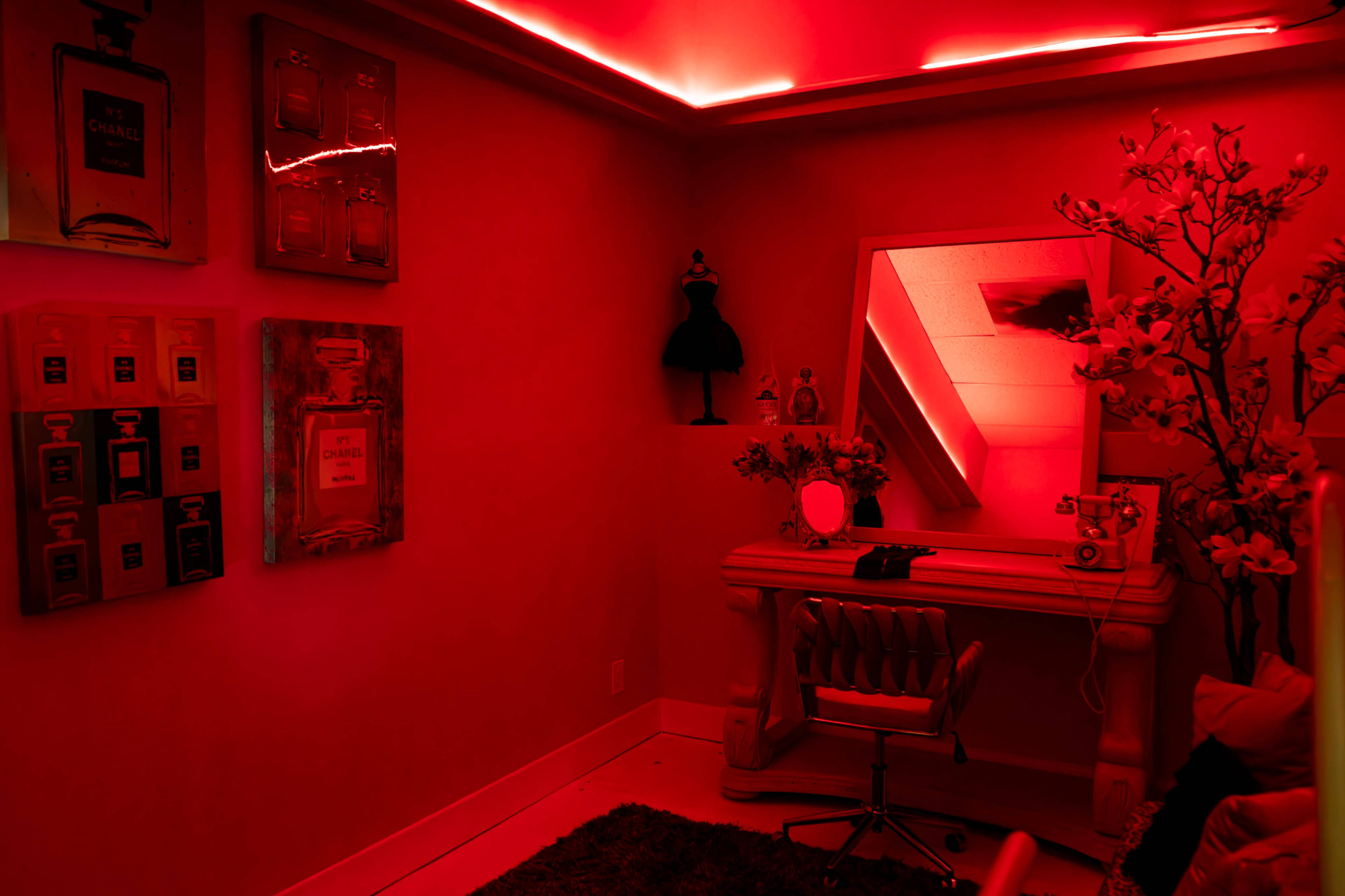 The room features red walls, illuminated by ambient lighting, with a mirrored vanity, decorative perfume posters, and a mannequin dress form.