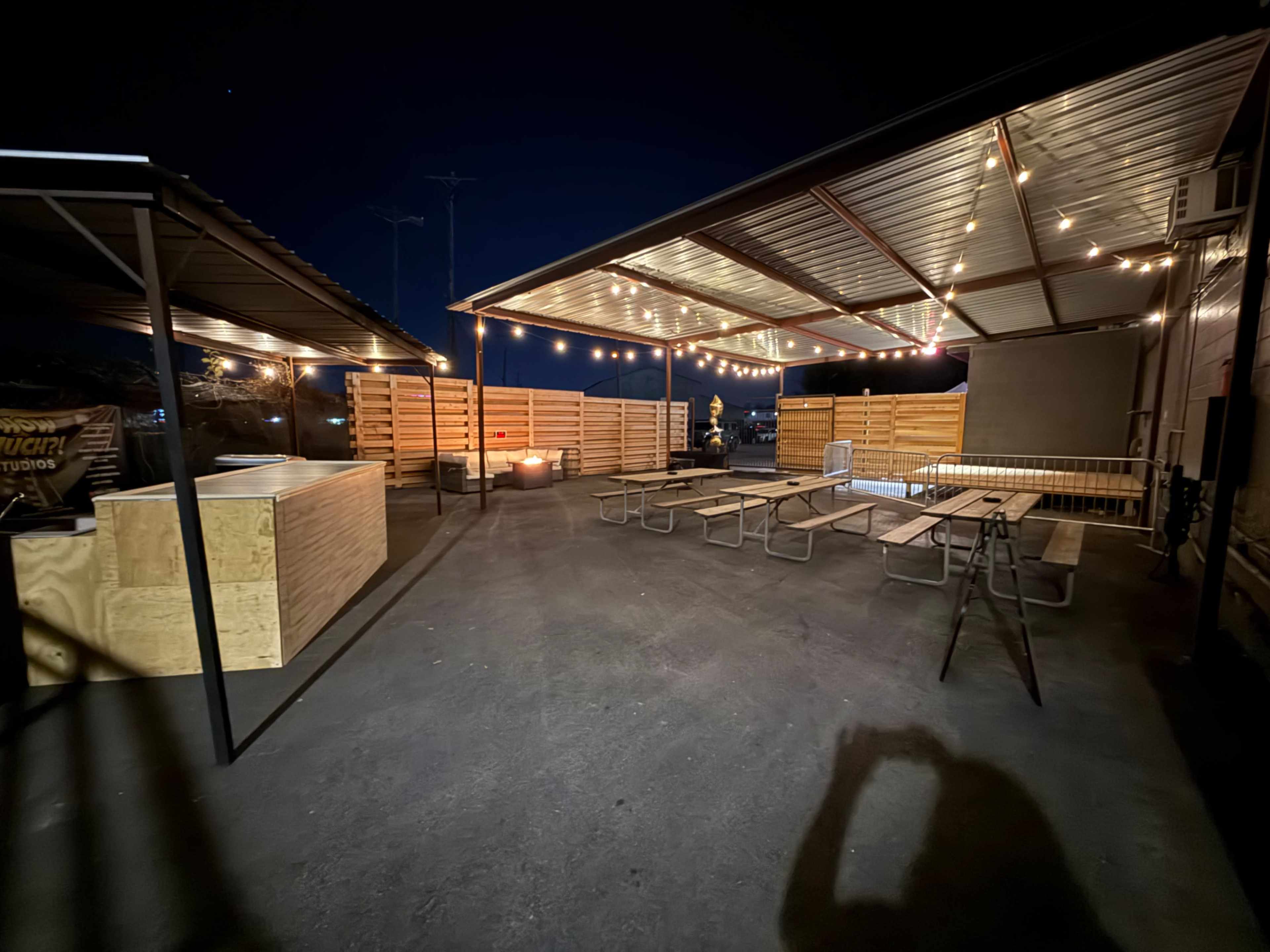 The image shows an outdoor seating area with wooden picnic tables and illuminated by string lights, enclosed by wooden partitions under a metal roof at night.