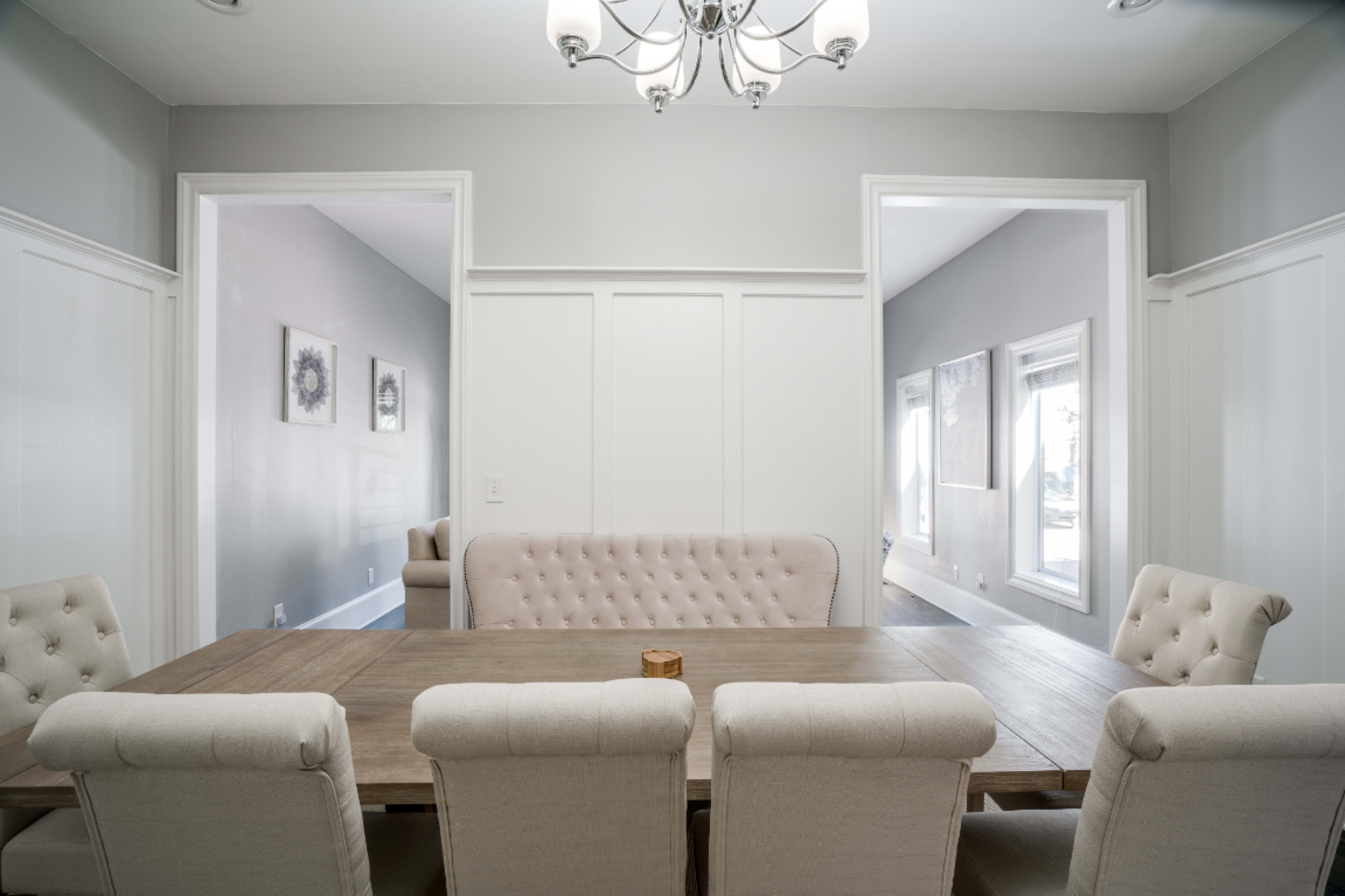 A light-colored dining room features a wooden table surrounded by upholstered chairs, with a tufted banquette against the wall and doorways leading to adjacent rooms.