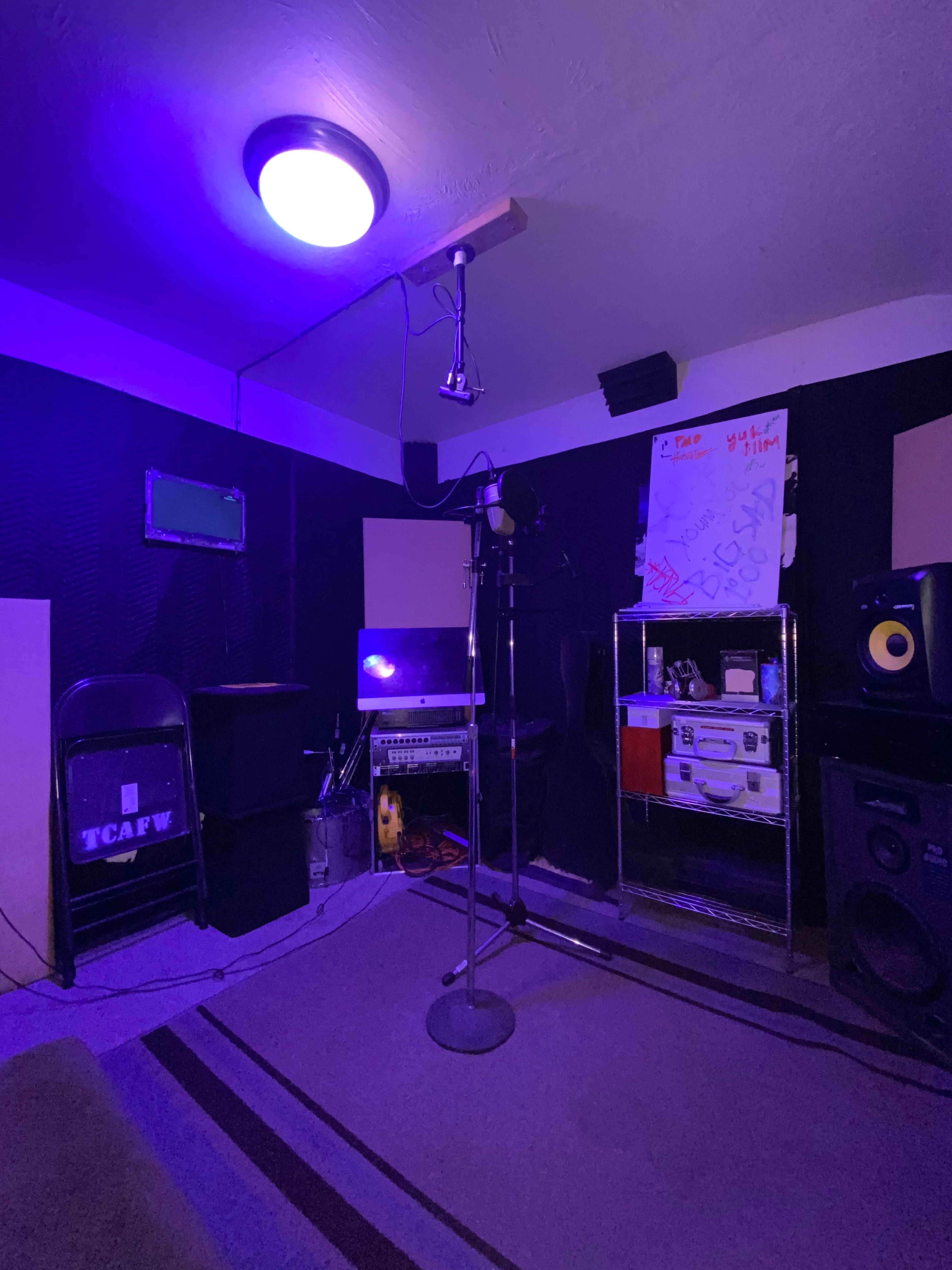 The image shows a recording studio with a microphone on a stand, shelves of equipment, and soundproofing panels surrounding the space.
