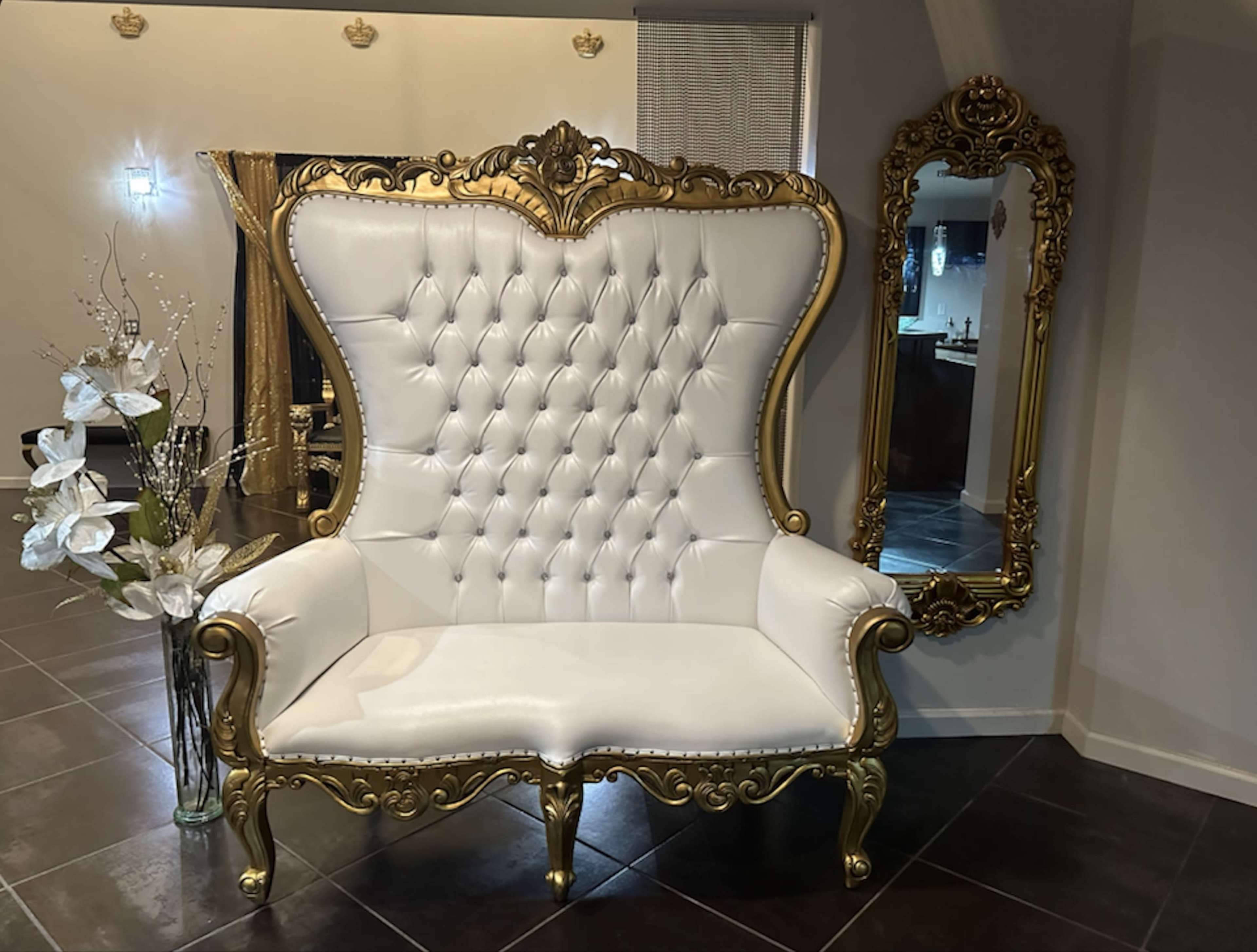 The image features a large, ornate white and gold tufted chair positioned next to an elaborate gold-framed mirror in a well-lit room.