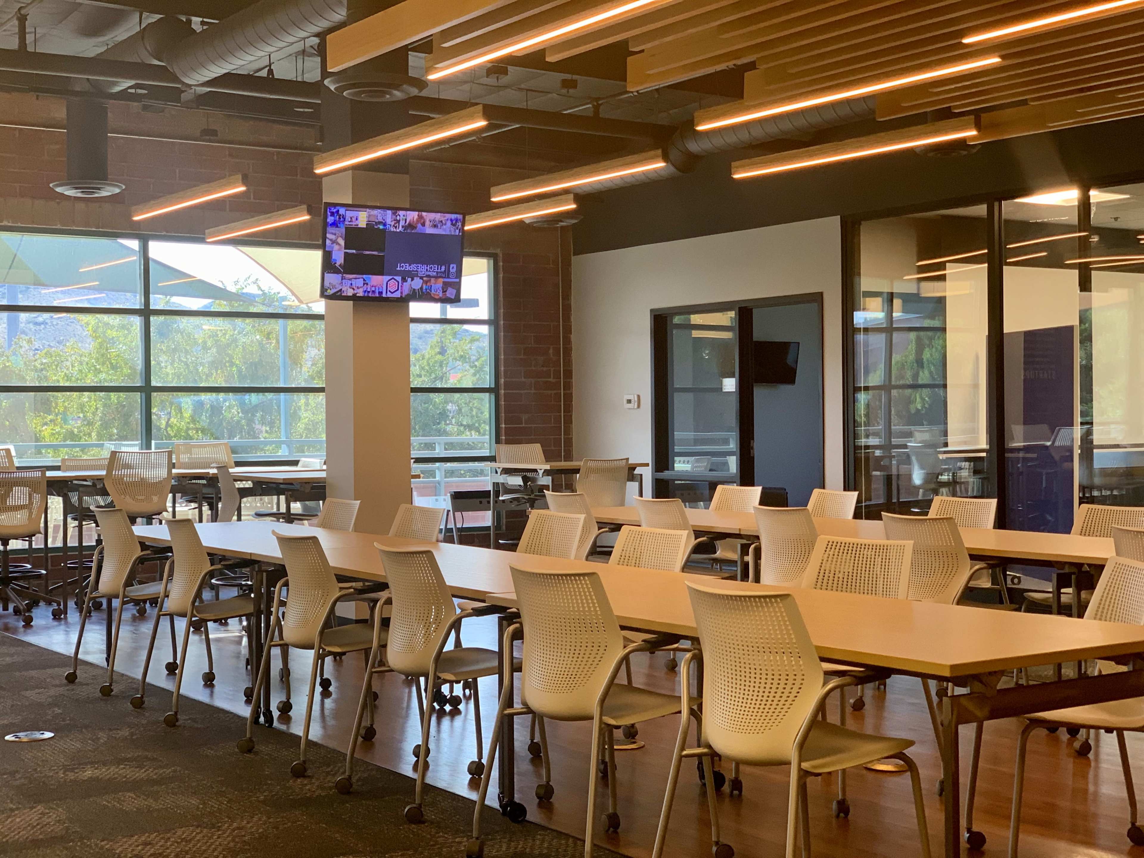 A modern conference room with multiple tables and chairs arranged for meetings, featuring large windows and a television screen mounted on the wall.