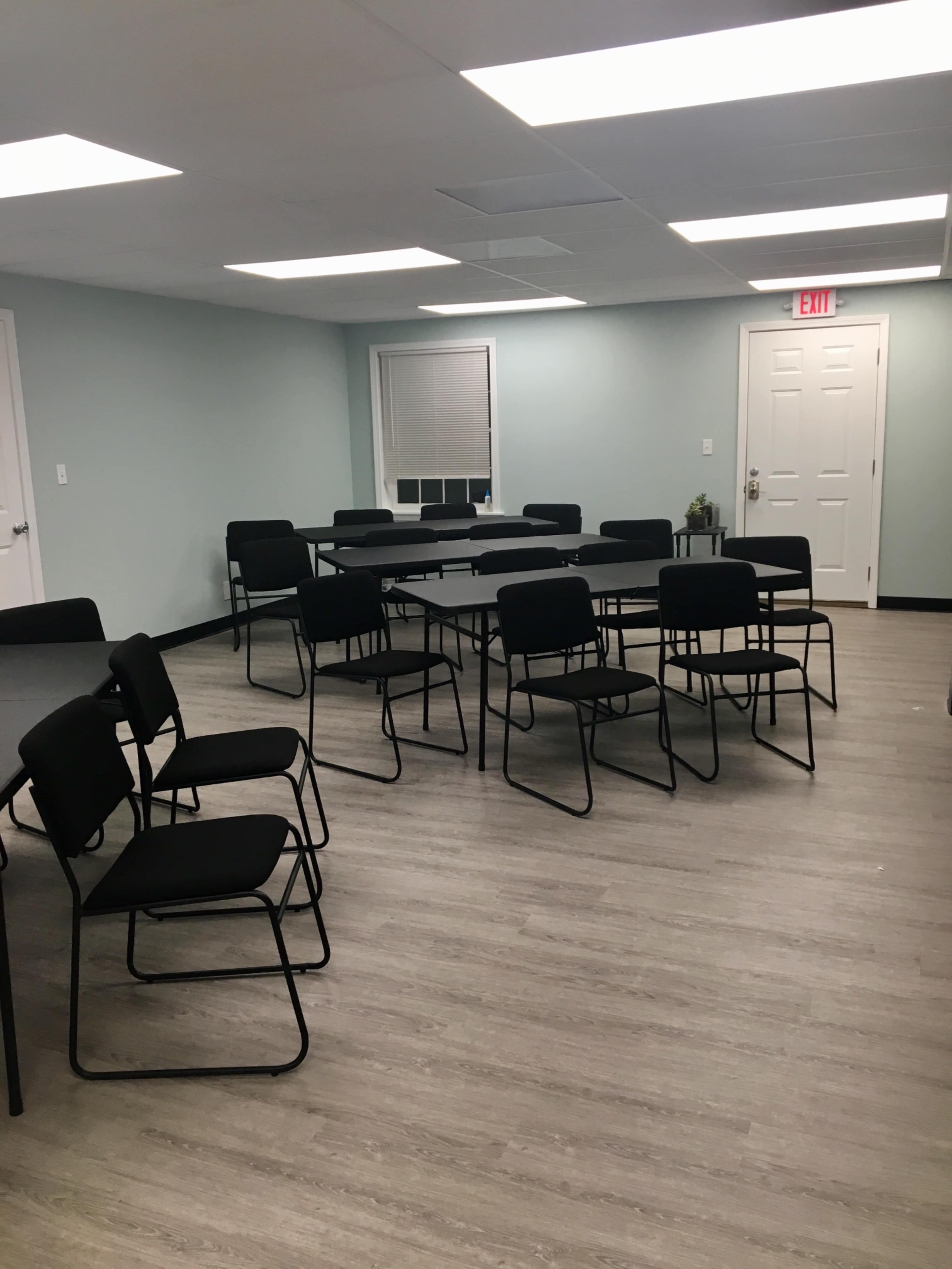 A meeting room is arranged with several black chairs and tables on a light wooden floor, with pale blue walls and ceiling lights.