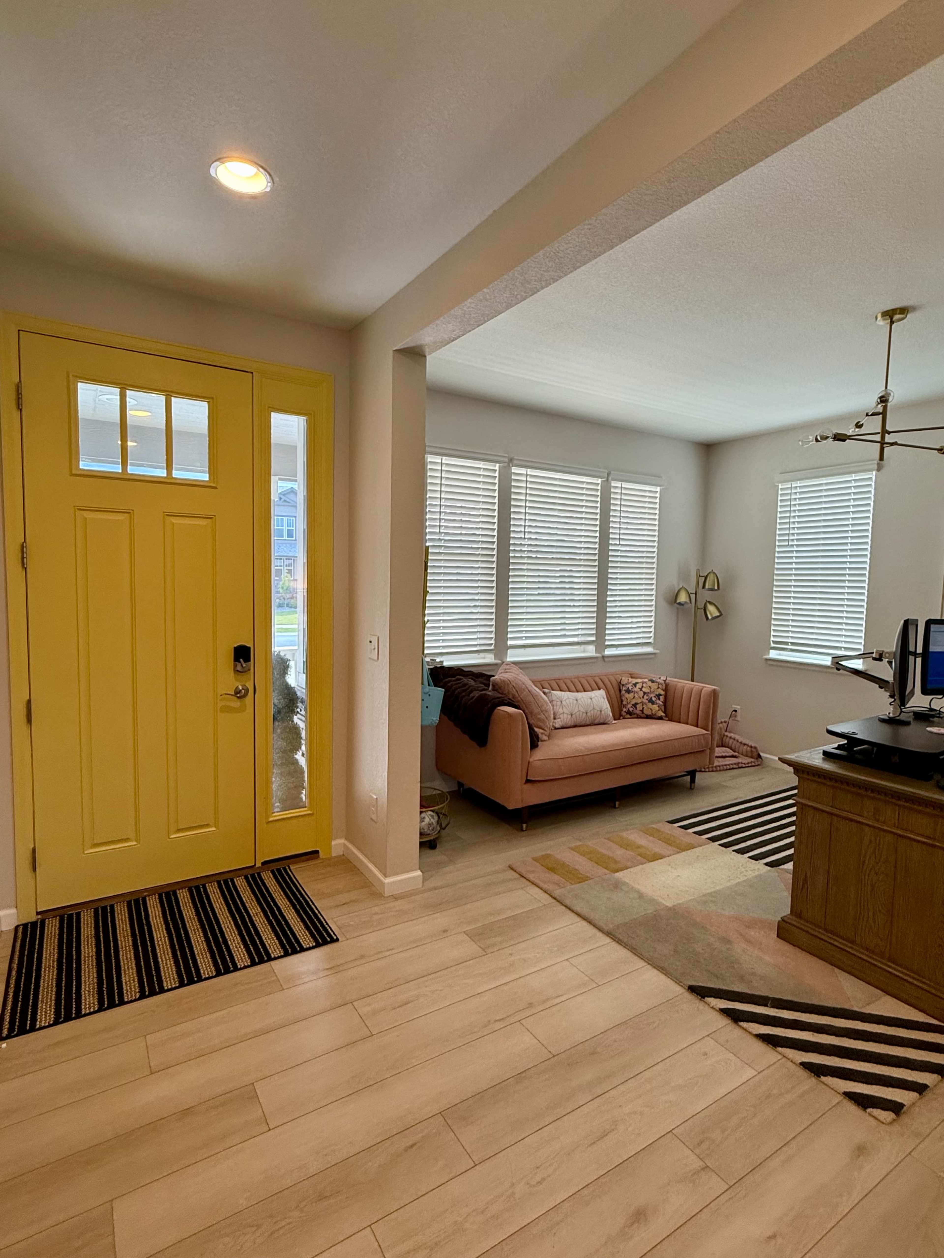 The image shows an entryway with a bright yellow front door, leading to a living area featuring a pink sofa and several windows with light filtering in.