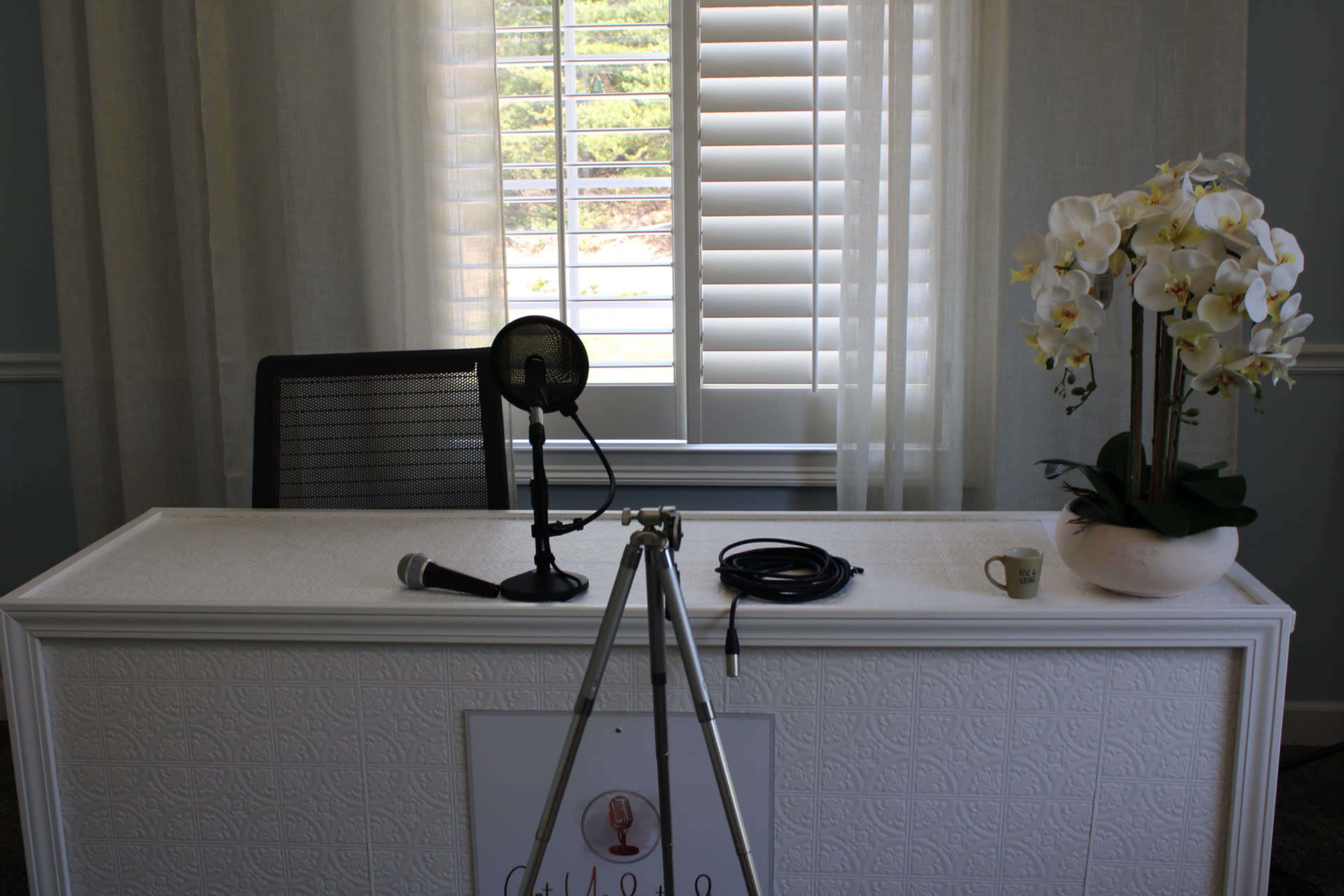 A simple office setup features a desk with a microphone, a tripod, and a potted orchid, positioned near a window with white blinds.