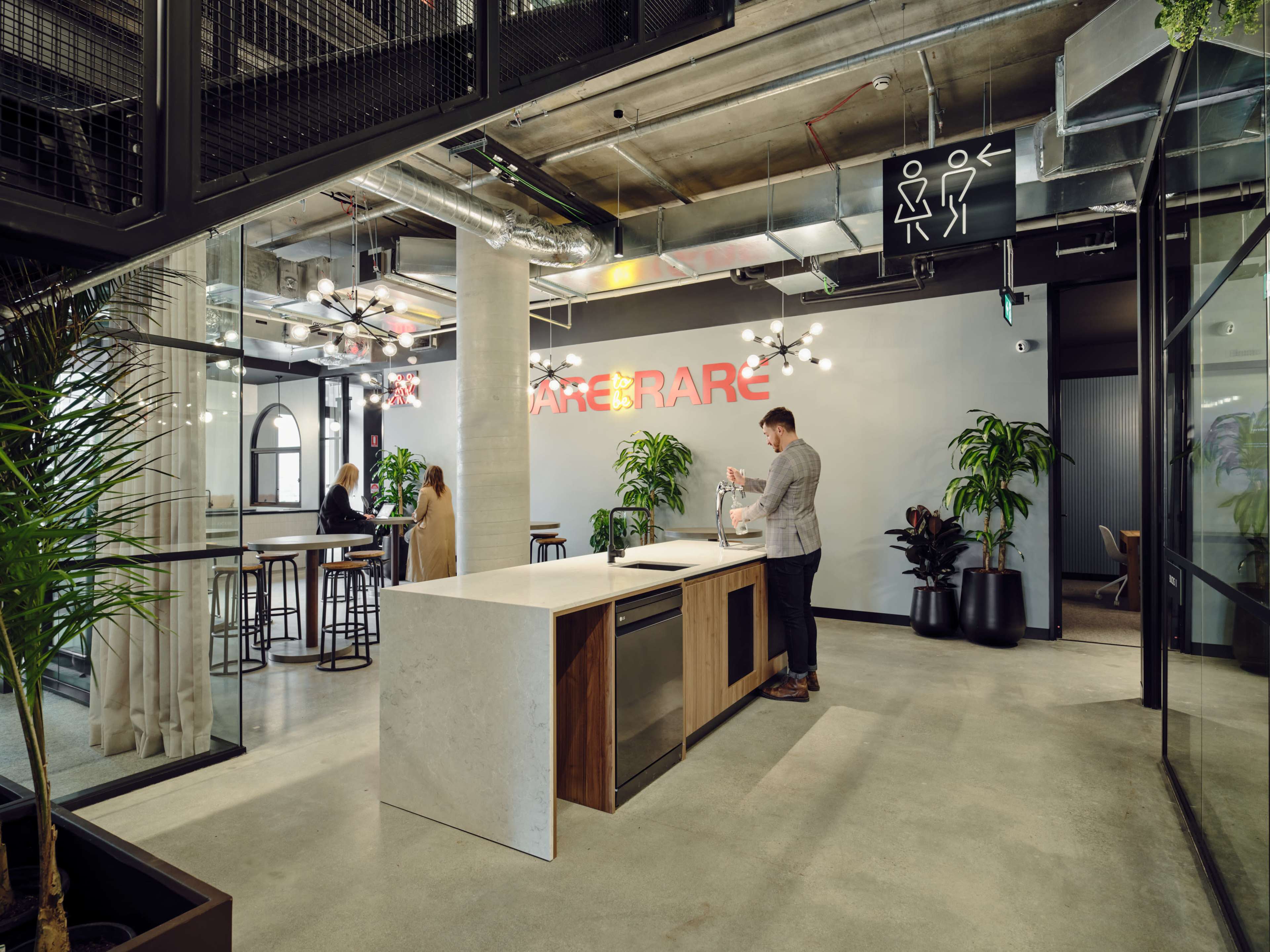 The image depicts a modern office reception area with large plants, stylish seating, and a minimalist countertop where a person is interacting with a visitor.