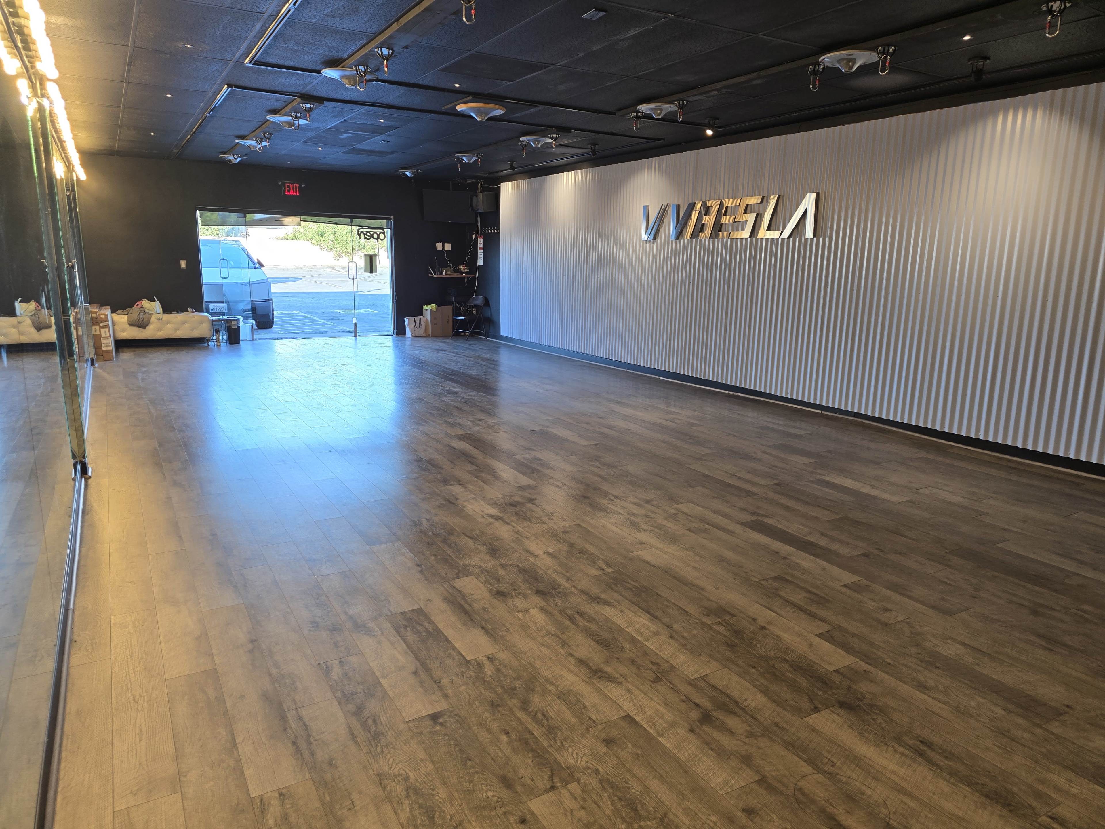 The image shows a spacious, empty room with wooden flooring and a wall featuring the brand name "VIVETESLA," illuminated by lights, and an entrance leading outside.