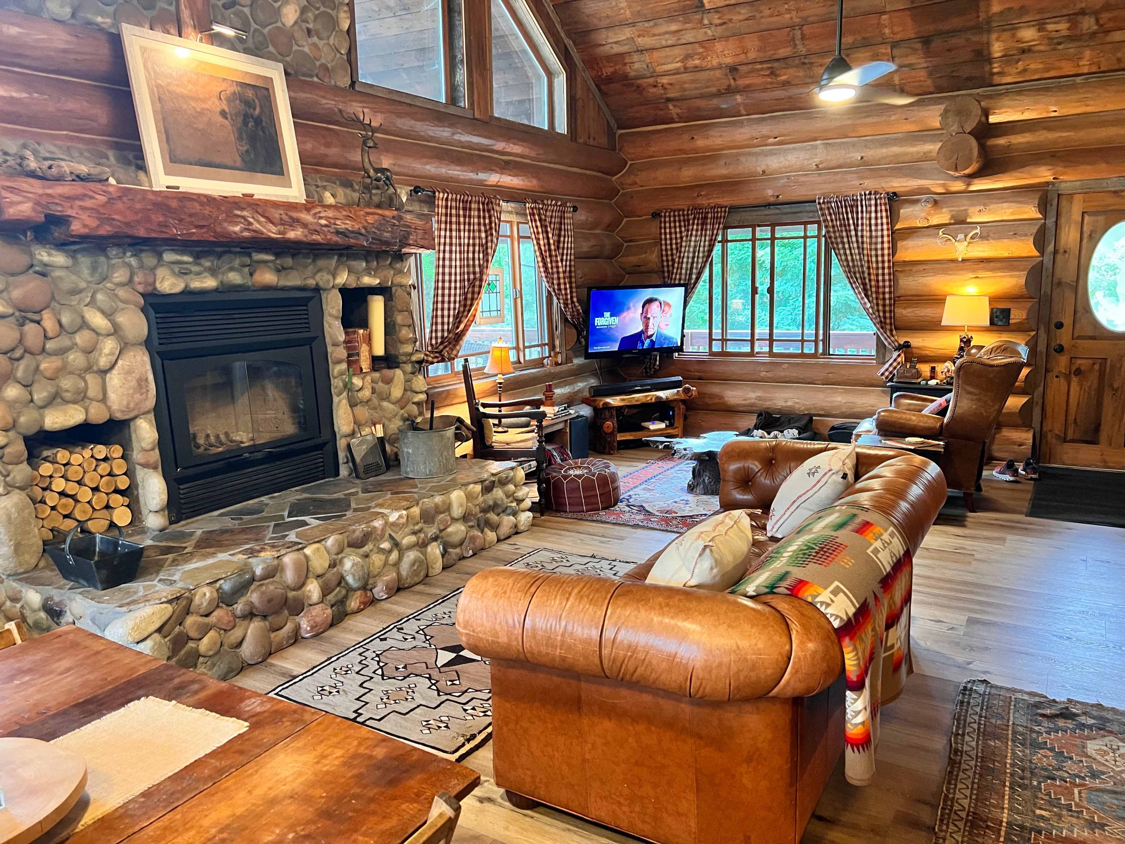 The interior of a log cabin features a stone fireplace, leather sofas, a wooden dining table, and large windows with natural light.