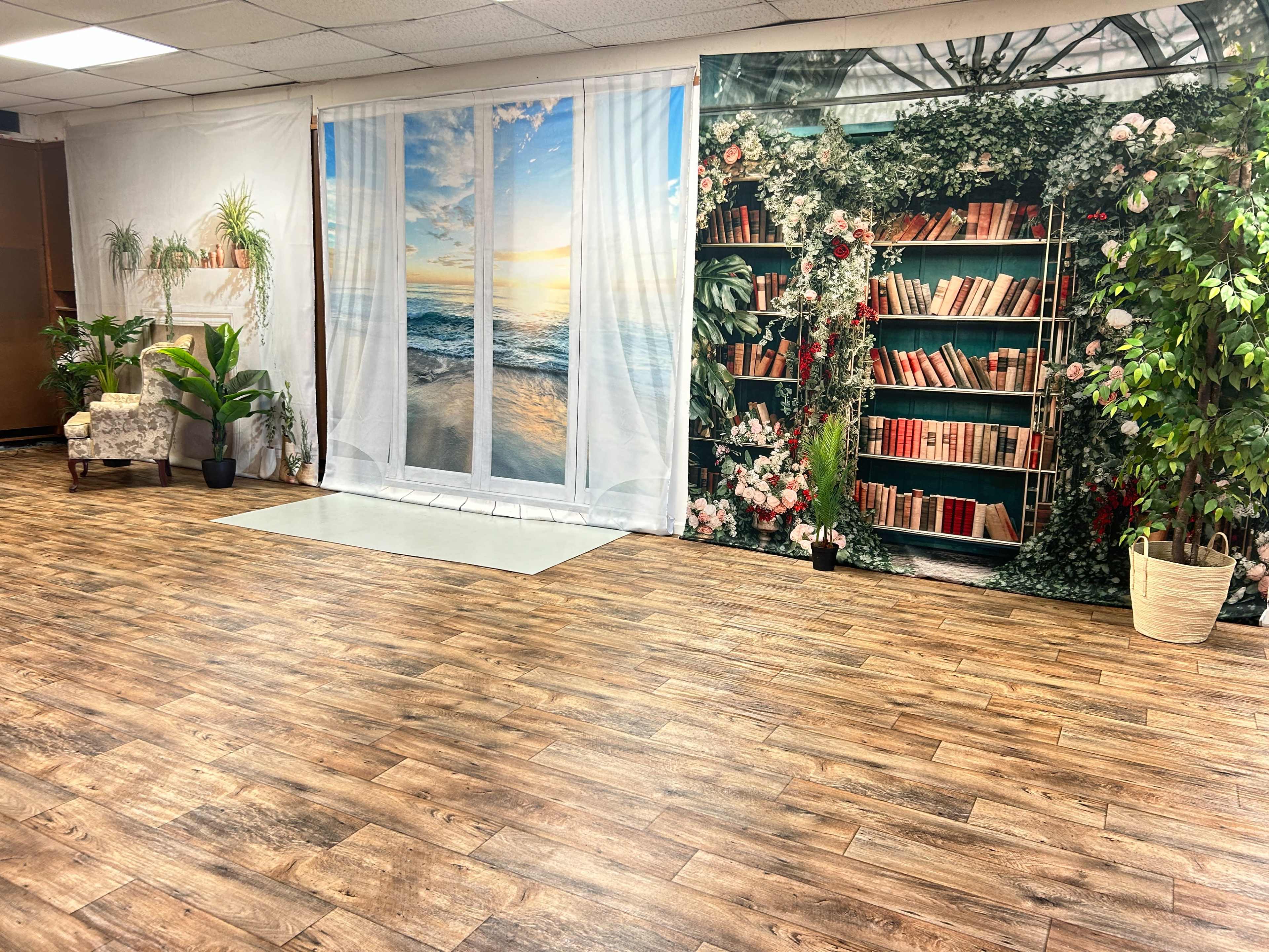 The image shows a room with a wooden floor, featuring a large backdrop of a bookshelf filled with books and a beach scene with a sunset through sheer curtains, along with potted plants and a chair in one corner.