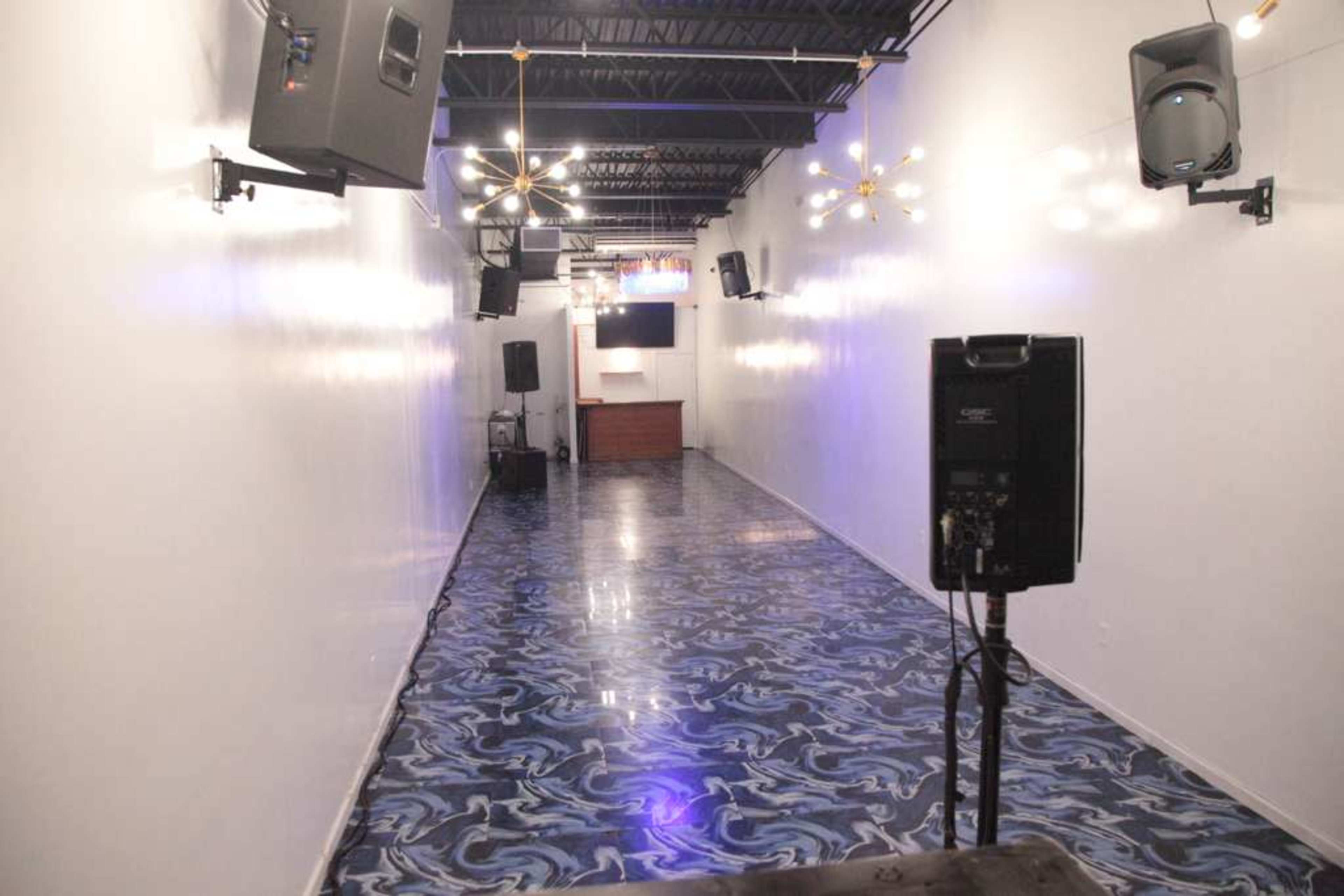The image shows a long, narrow room with a shiny blue patterned floor and white walls, featuring speaker systems mounted on the walls and a small stage or counter at the far end.