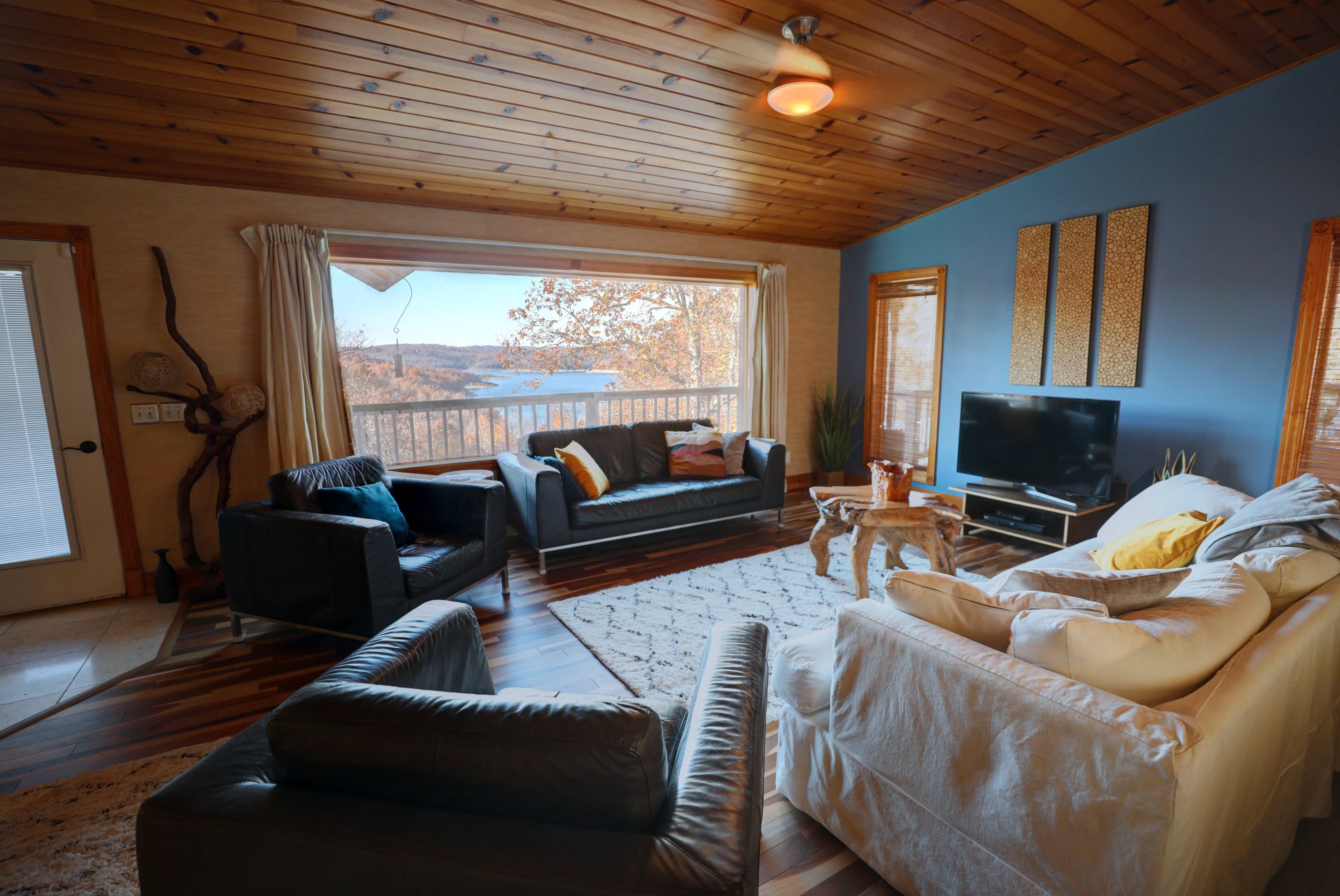 The room features a cozy living area with a view of the outdoors, furnished with a mix of dark and light sofas, a wooden coffee table, and a large window letting in natural light.
