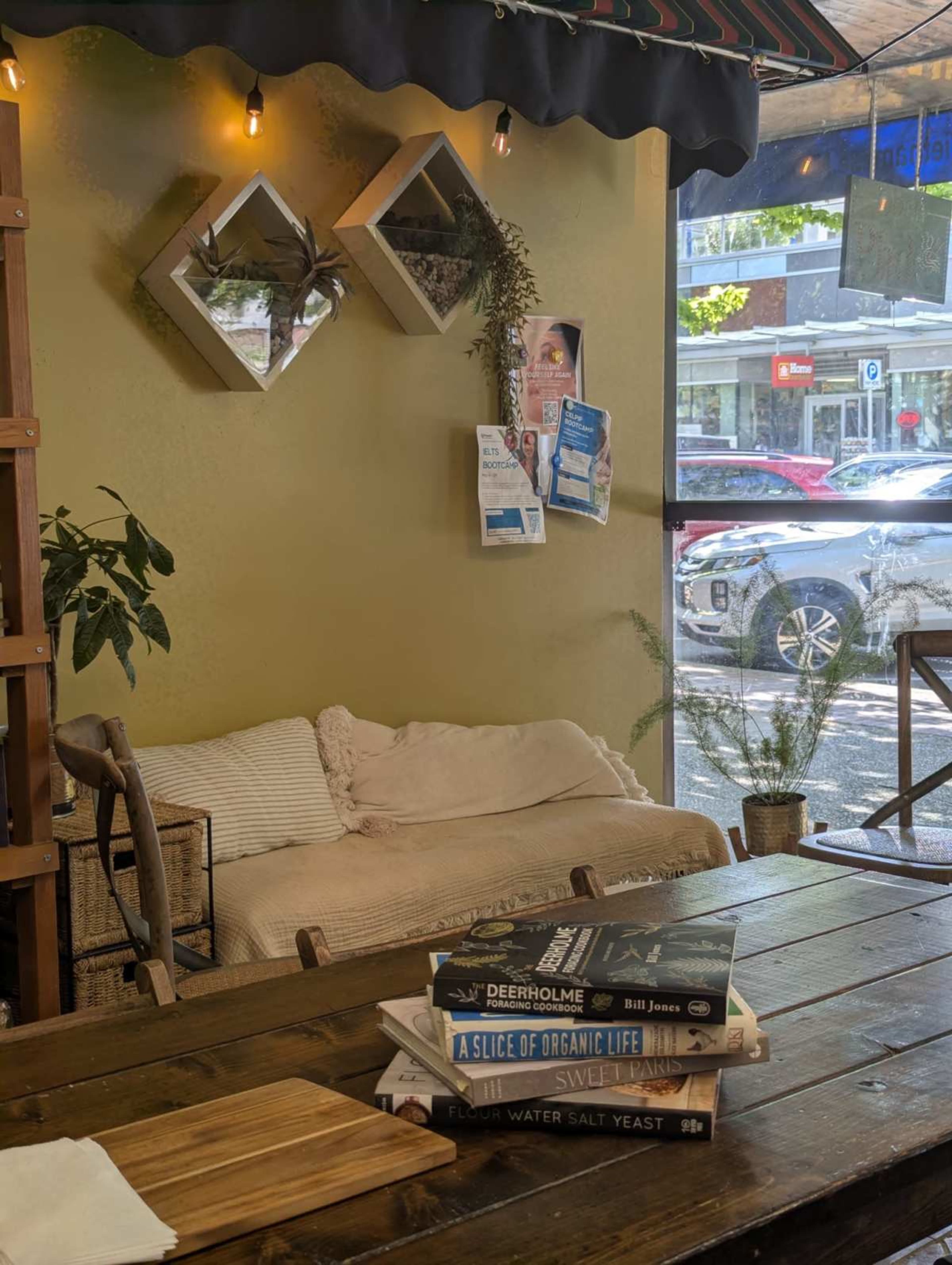 The image shows a cozy café interior featuring a couch and a wooden table stacked with books, while large windows overlook a busy street outside.