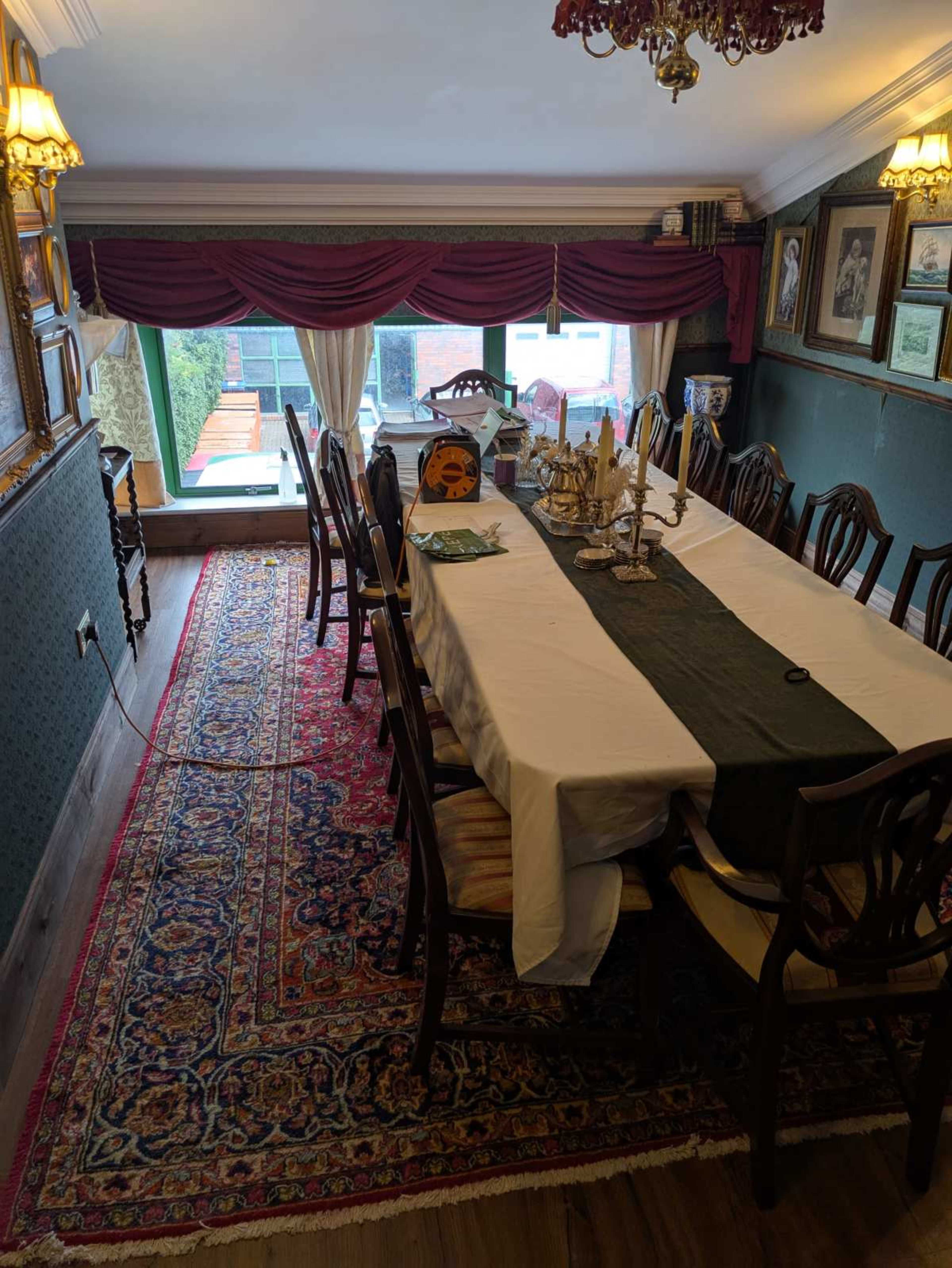 A long dining table is set with a green runner in a room decorated with framed pictures and a large window displaying greenery outside.