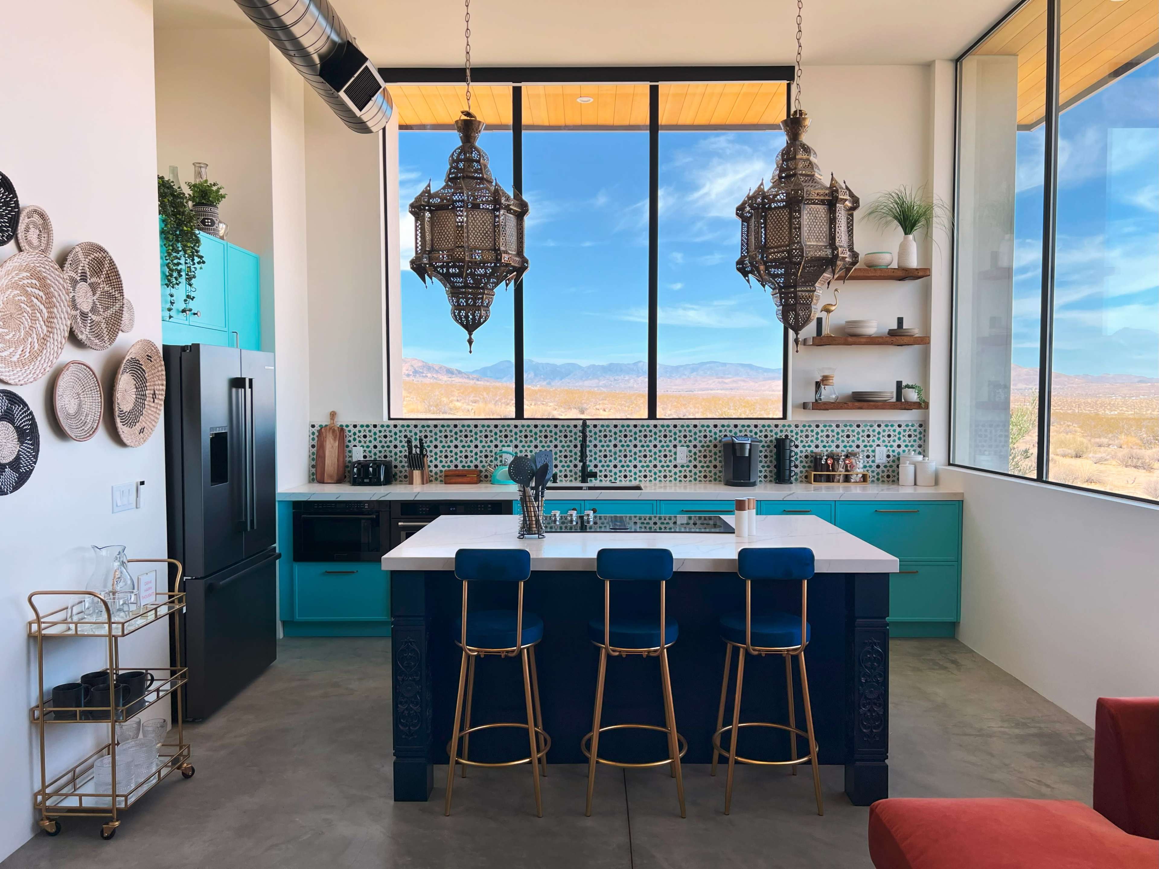 The kitchen features a modern design with turquoise cabinetry, a large island, and pendant lighting overlooking a desert landscape through large windows.