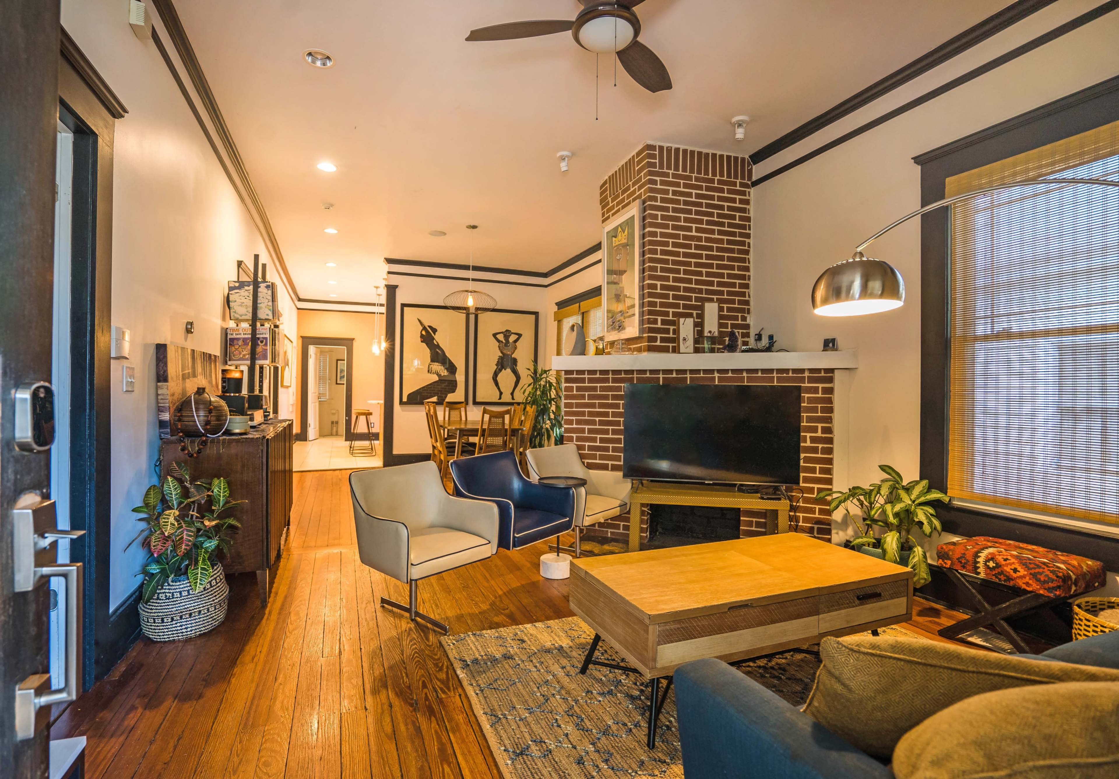 A cozy living room featuring wooden floors, a brick fireplace, modern furniture, and various plants.