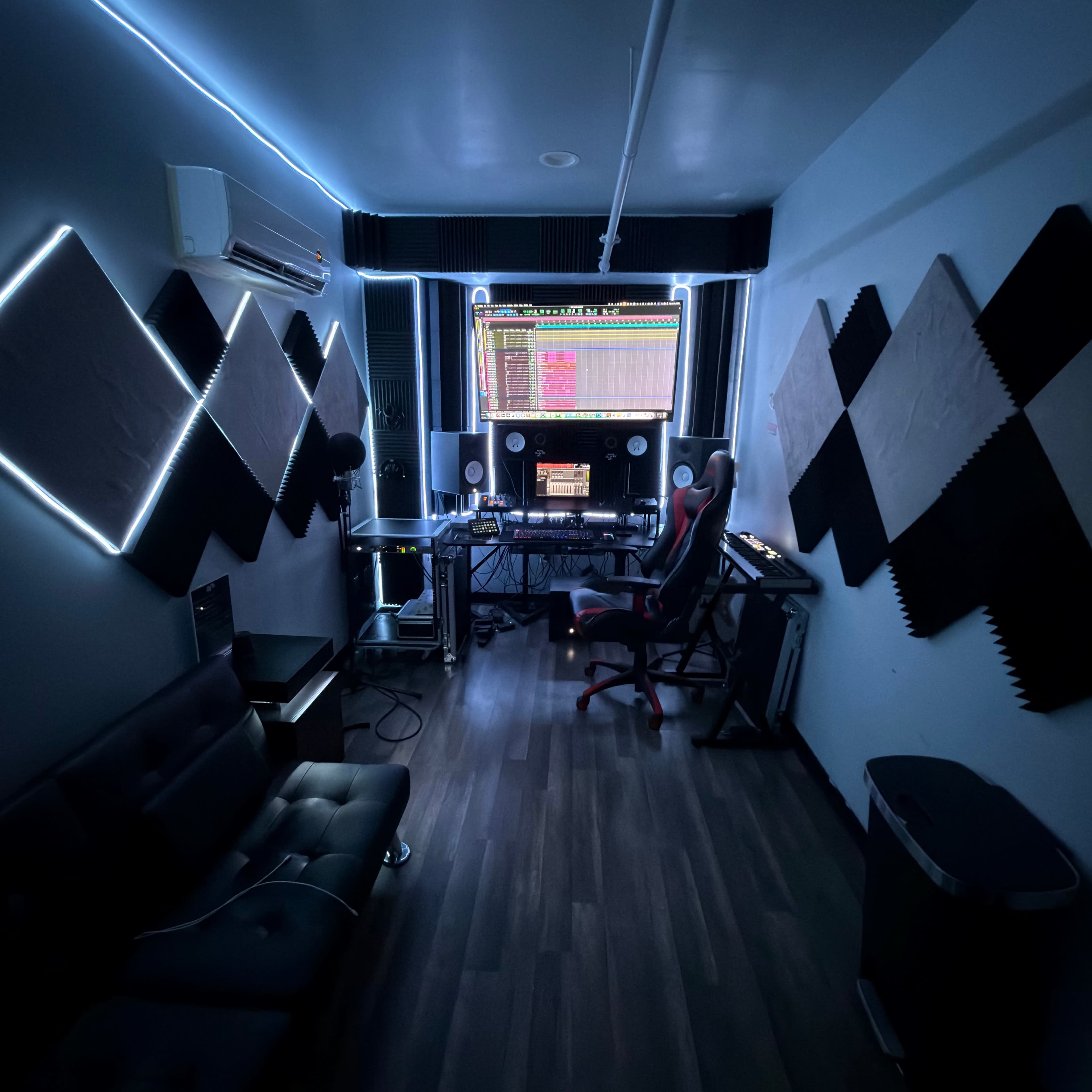 The image shows a modern music production studio featuring soundproof walls, a large computer monitor displaying audio software, and a sleek chair positioned at a workstation.