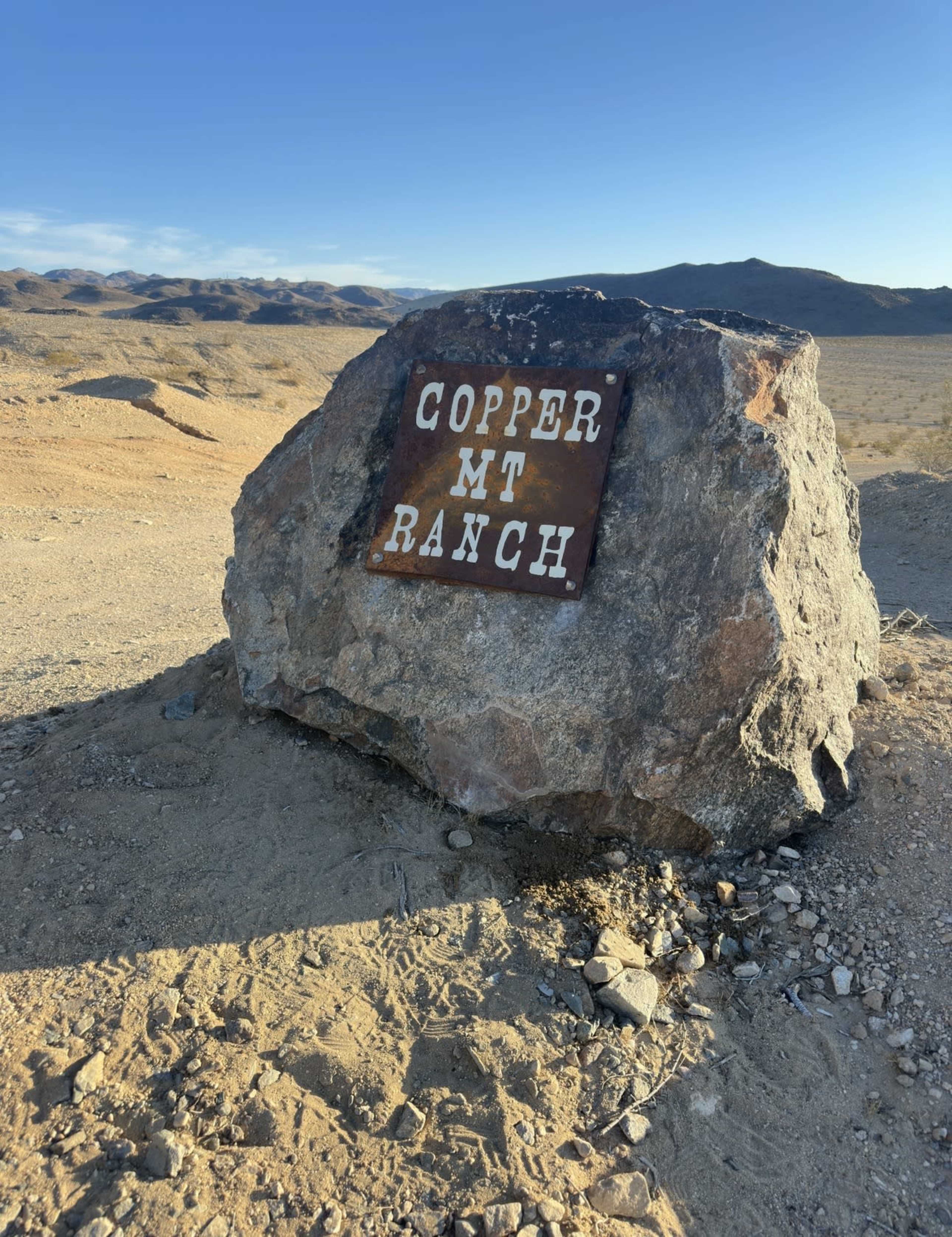A large rock with a sign reading "COPPER MT RANCH" is situated in a barren desert landscape under a clear sky.