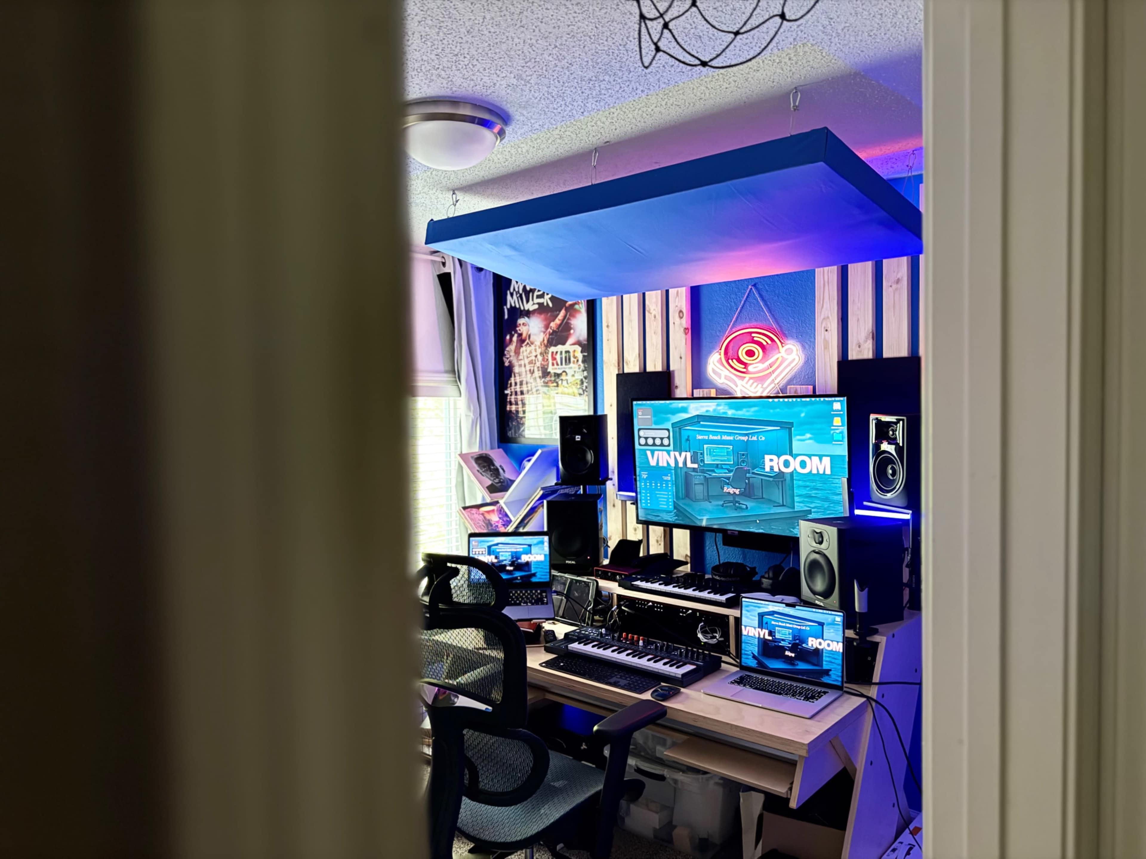 The image shows a colorful music production studio with multiple monitors, a keyboard, and LED lighting, viewed through a doorway.