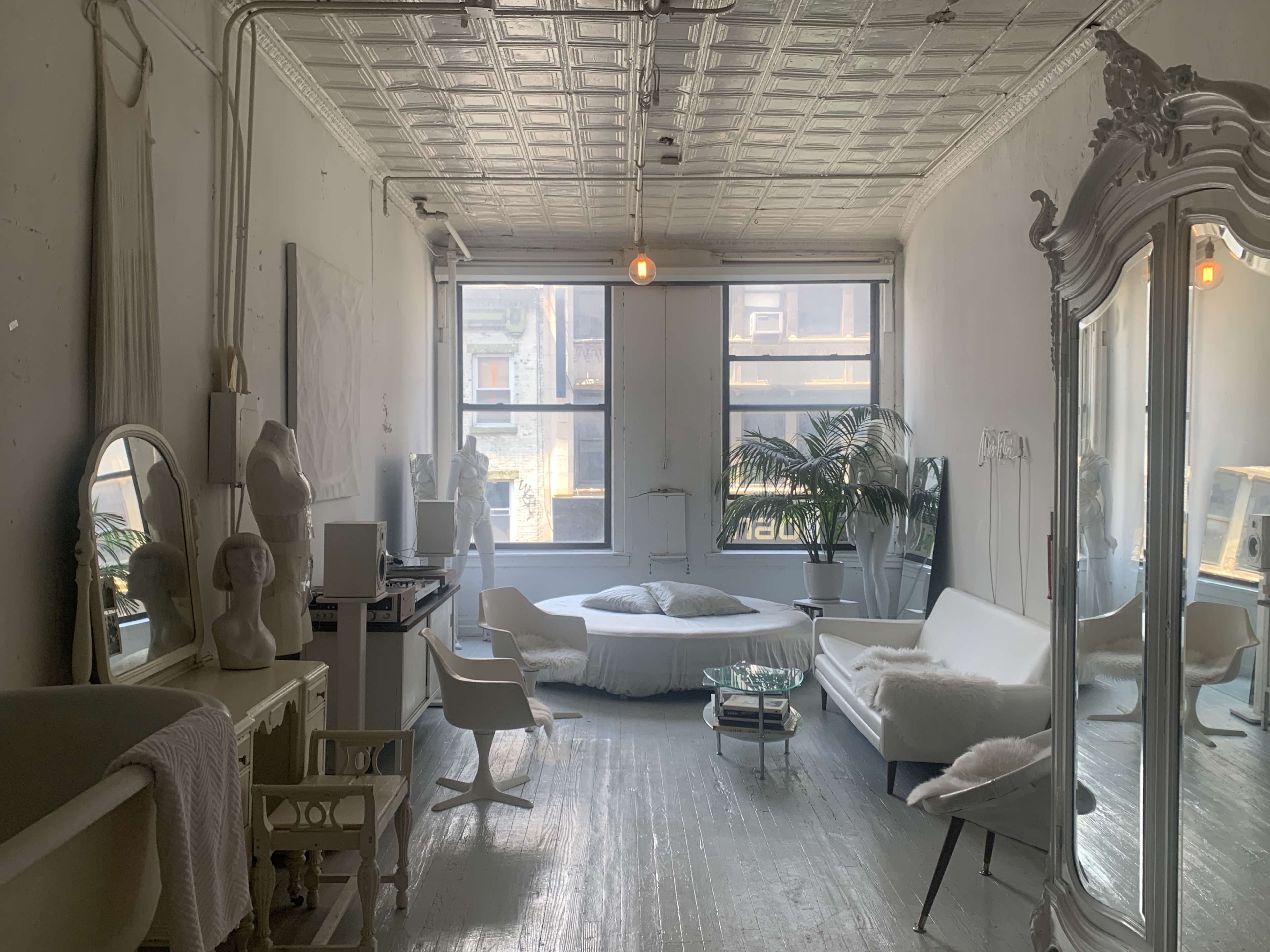 The image shows a bright, minimalist room with large windows, featuring a white sofa, a bed, a mirrored wardrobe, and various decorative elements.