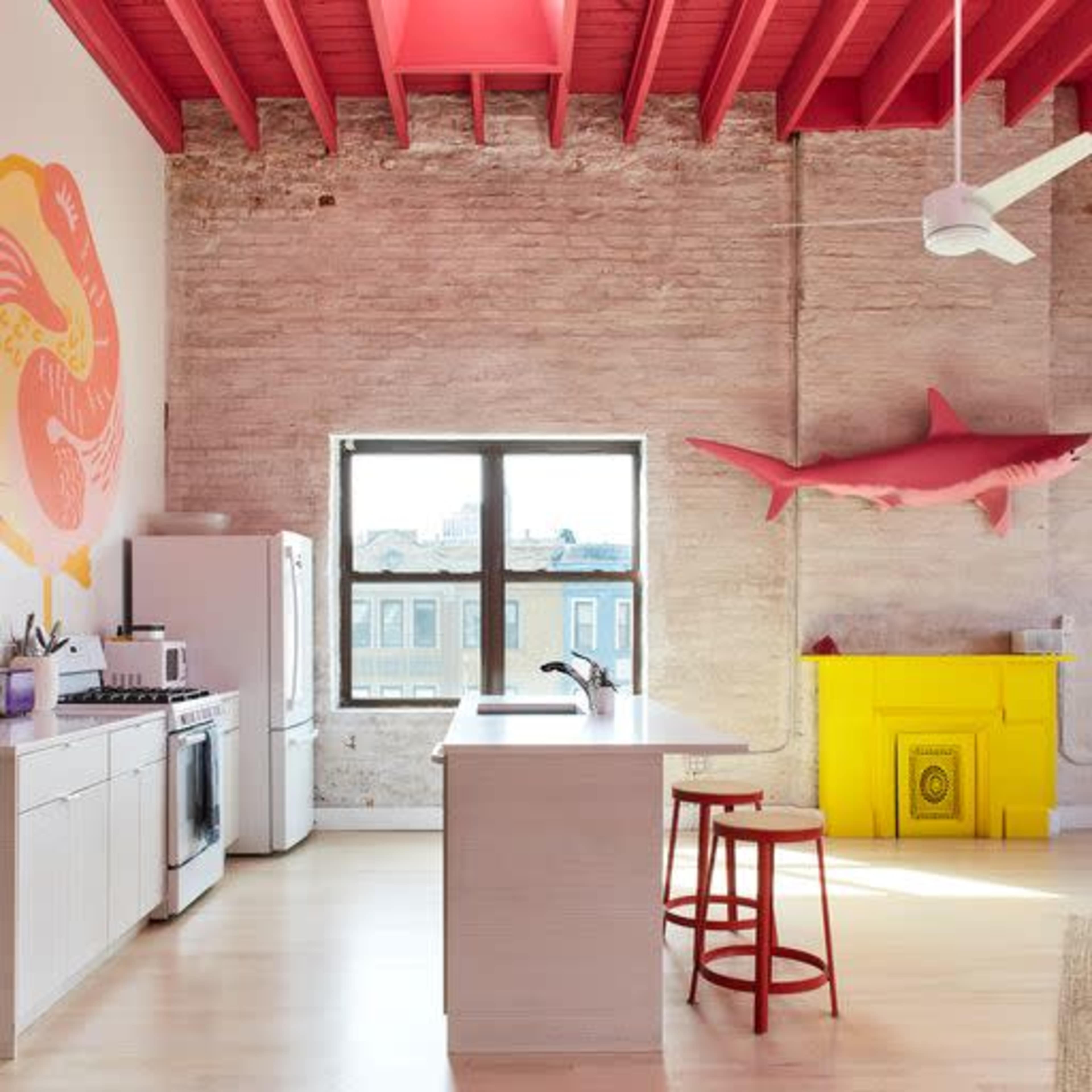 The image shows a modern kitchen with white cabinetry, a large island, and a window revealing an urban view, complemented by bright red accents on the ceiling and walls.