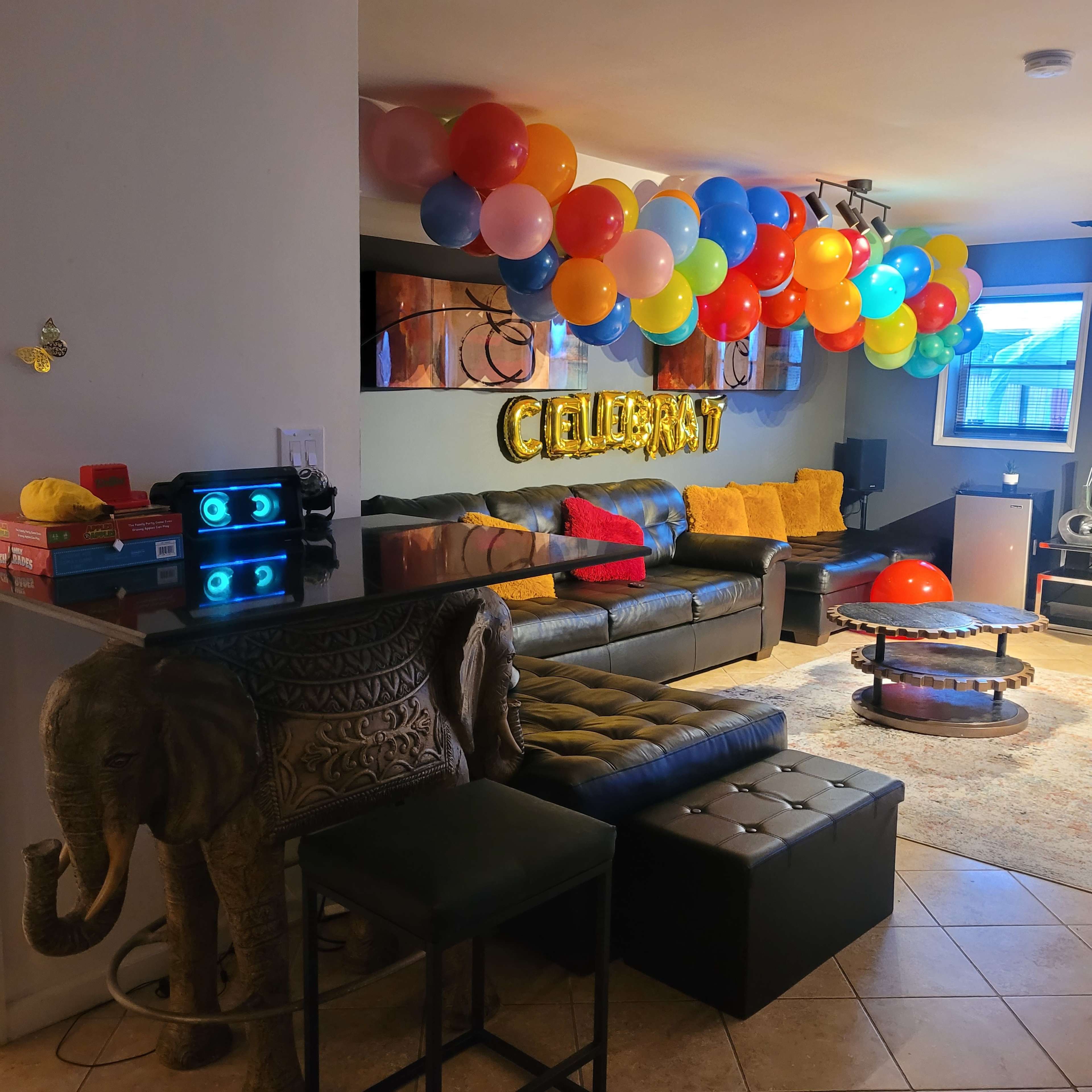 The living room features a black leather sofa, colorful balloons overhead, and an elephant-shaped side table.
