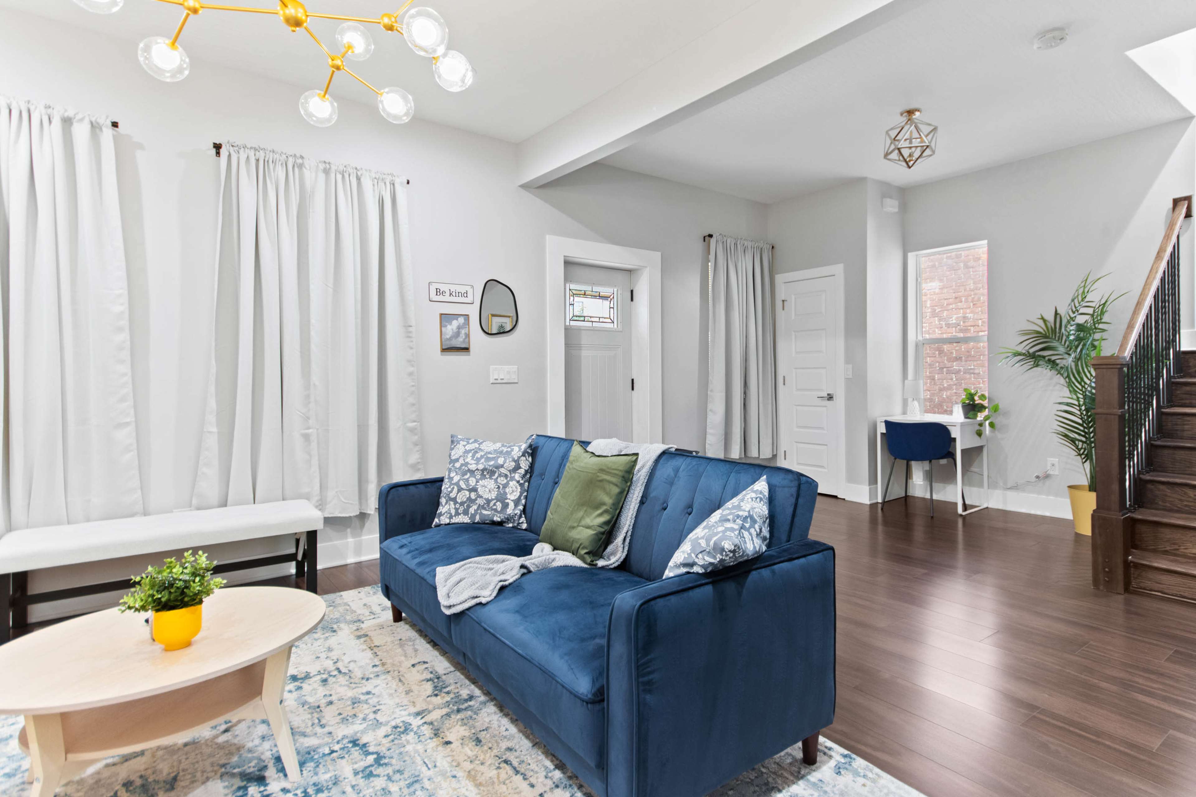 A modern living room features a blue sofa with decorative pillows, a circular coffee table, and large windows adorned with white curtains.