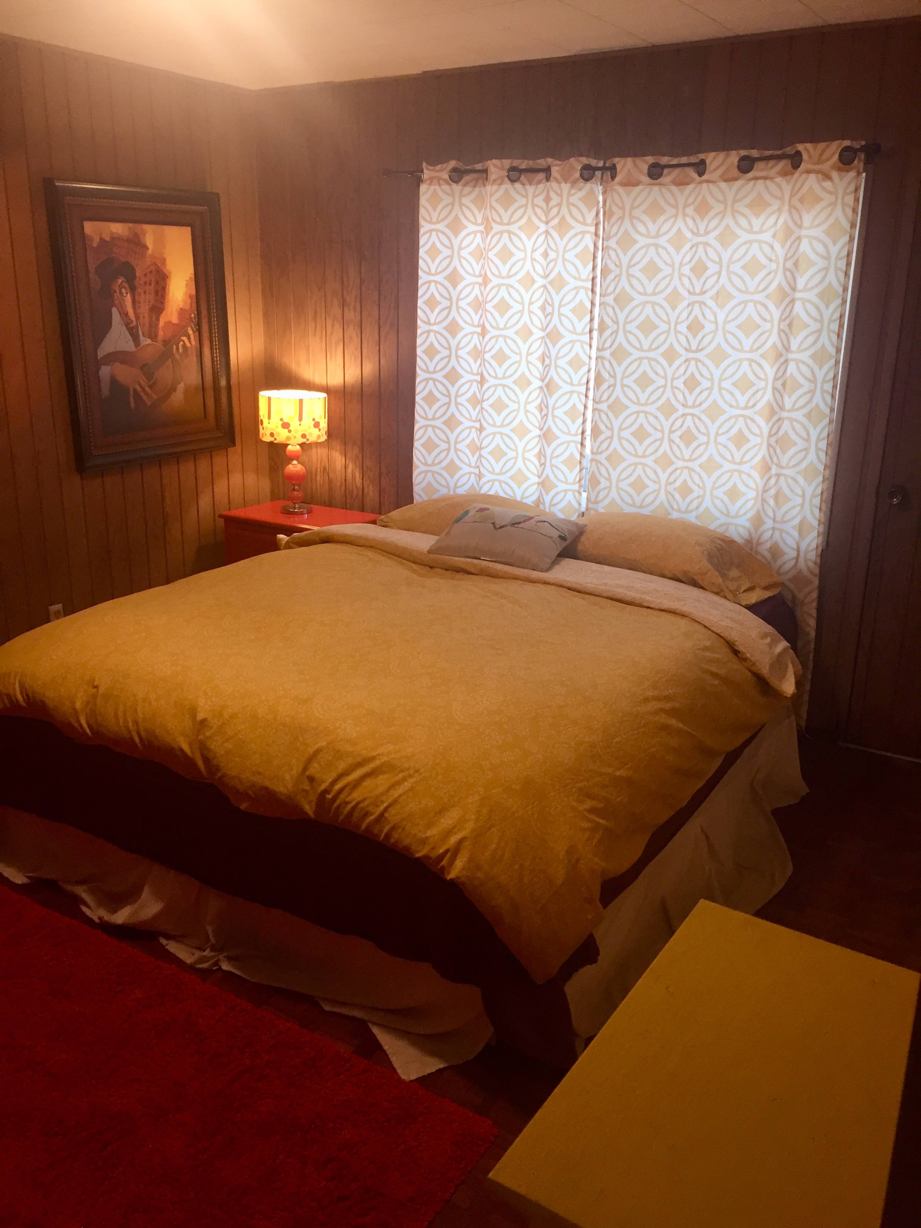 The image shows a cozy bedroom with a large bed, patterned curtains, a lamp, and a yellow nightstand.