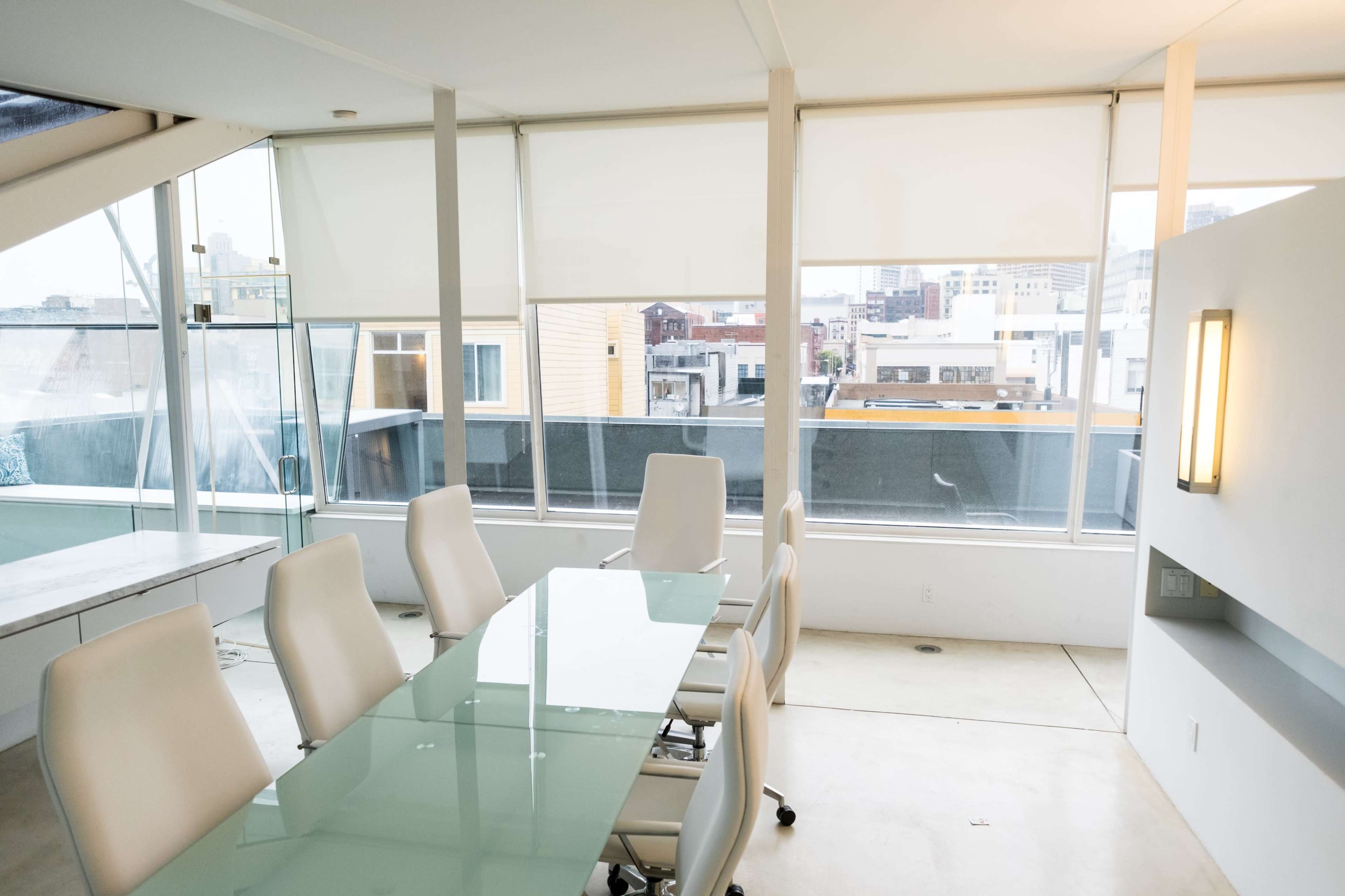 A conference room features a glass table surrounded by white chairs, with large windows offering a city view.